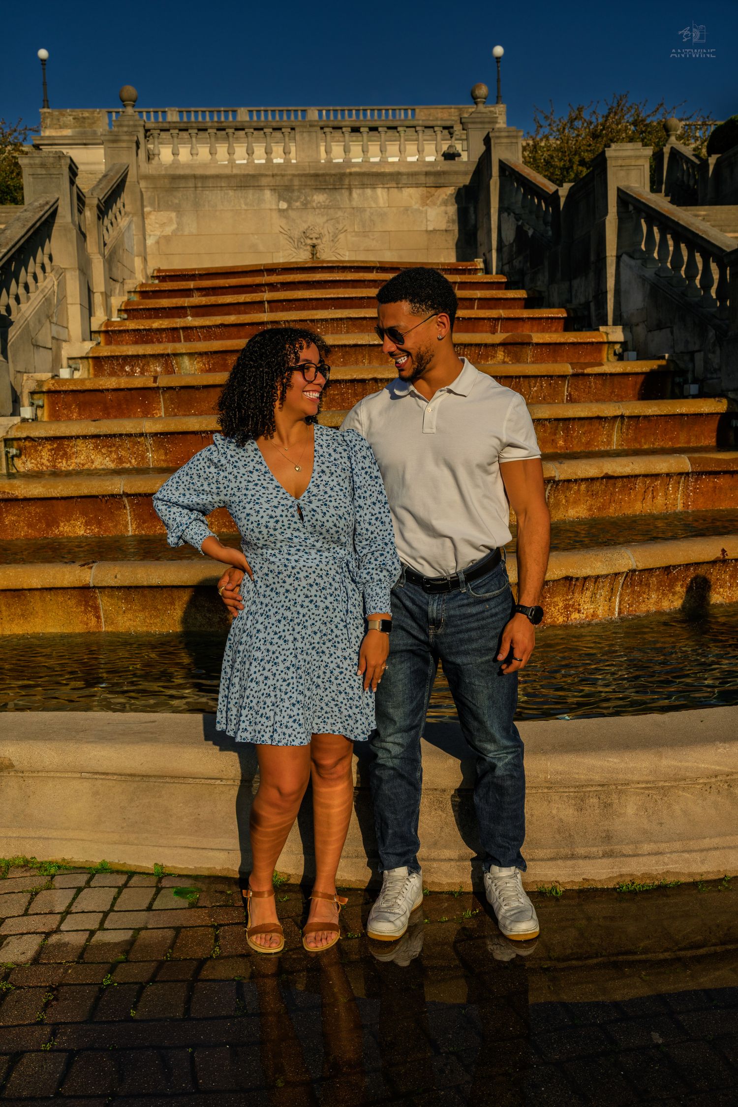 Couple smiles at each other standing before a decorative fountain and stone staircase at golden hour.