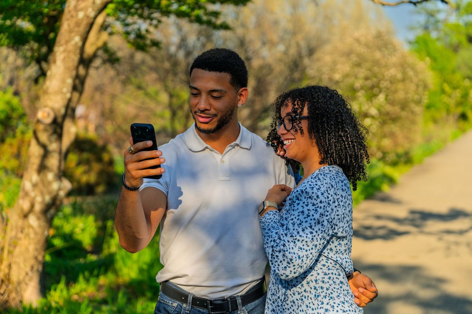 Happy couple looking at smartphone together on sunny park path, man in white polo embracing curly-haired woman.