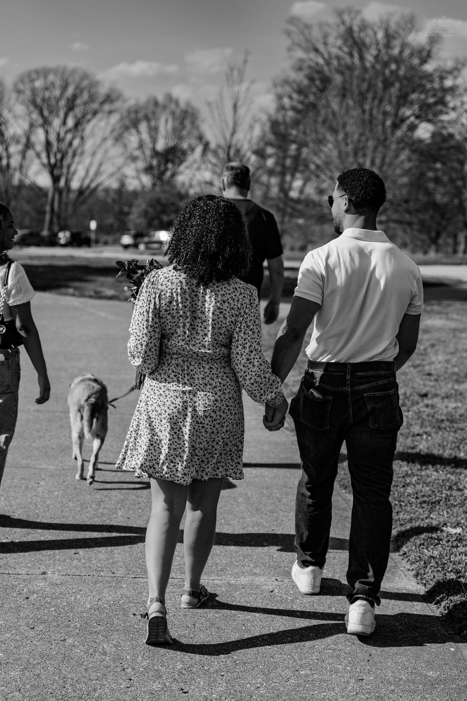 Black and white photo of couple holding hands walking in park with dog on leash nearby.