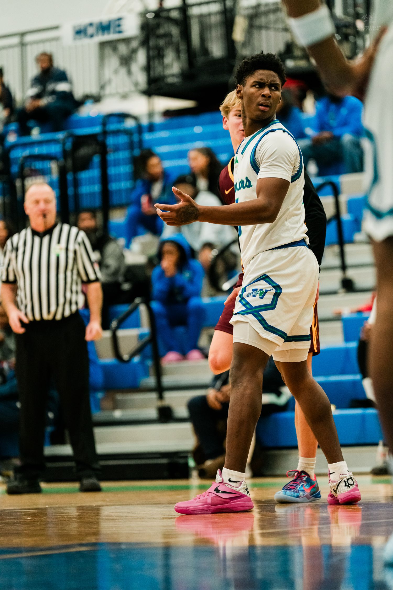 High school basketball player in white uniform gestures to teammates during competitive game with referee nearby.