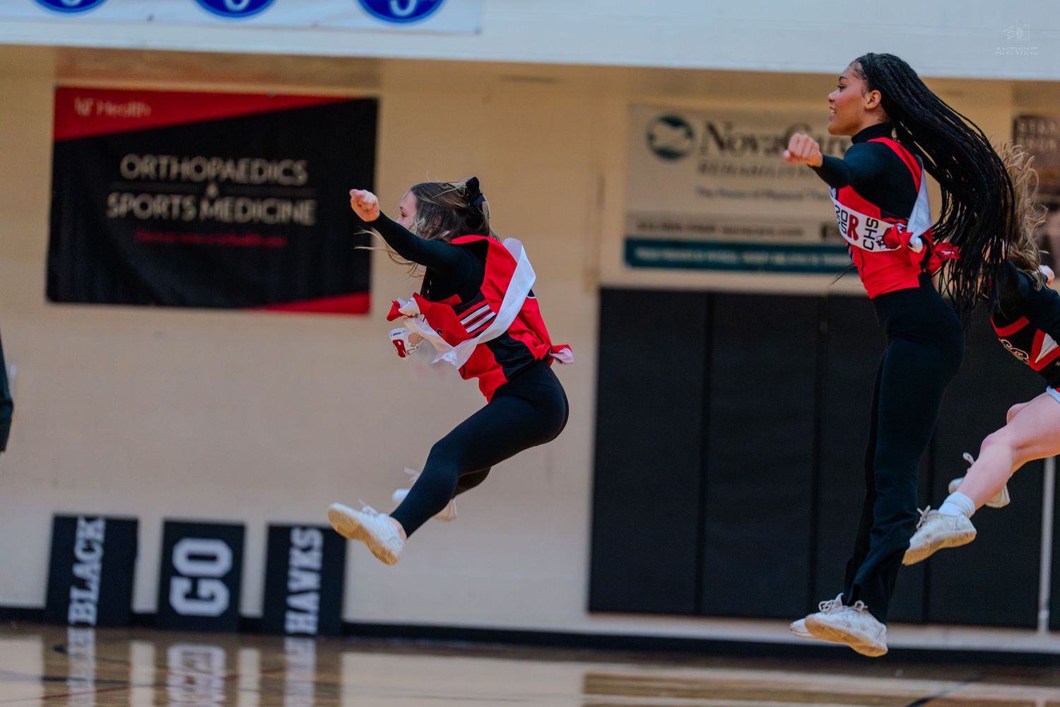 Two high school cheerleaders leap into the air during an energetic performance on a basketball court.