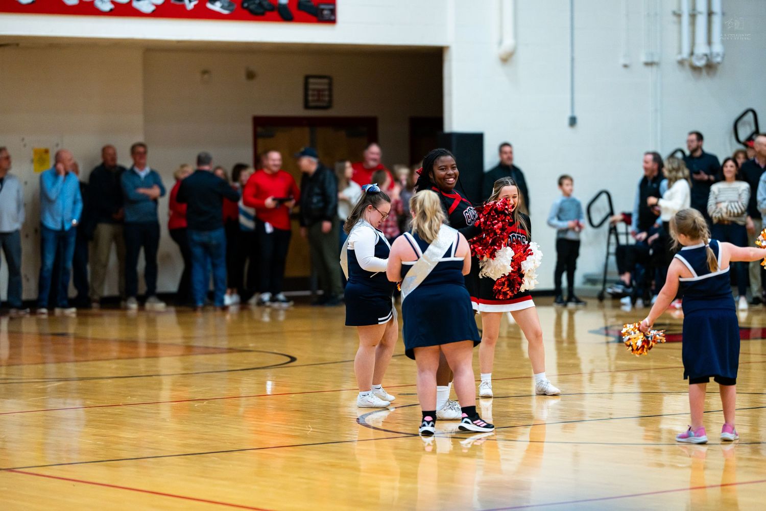 Youth cheerleaders in navy uniforms interact with college cheerleader on basketball court during game event.