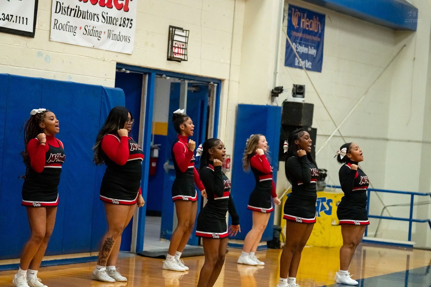 High school cheerleaders in red and black uniforms stand in a line cheering during a basketball game.