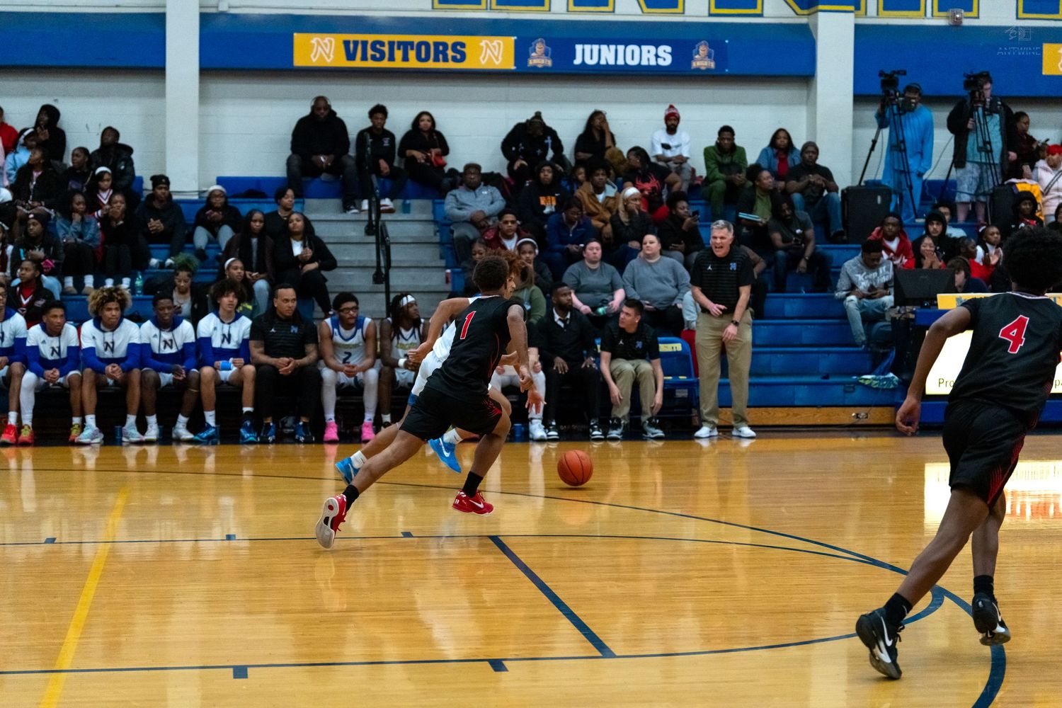 Basketball player in black dribbles near the baseline during a high school game with fans watching.