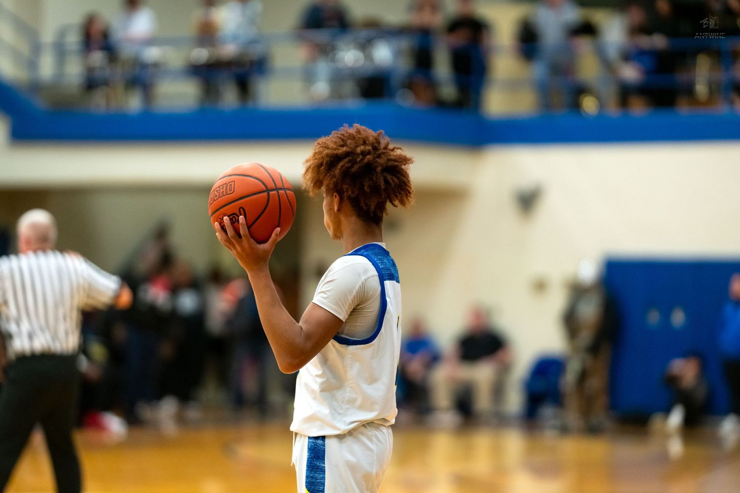 Young basketball player with curly hair holds a basketball during a high school indoor gymnasium game.
