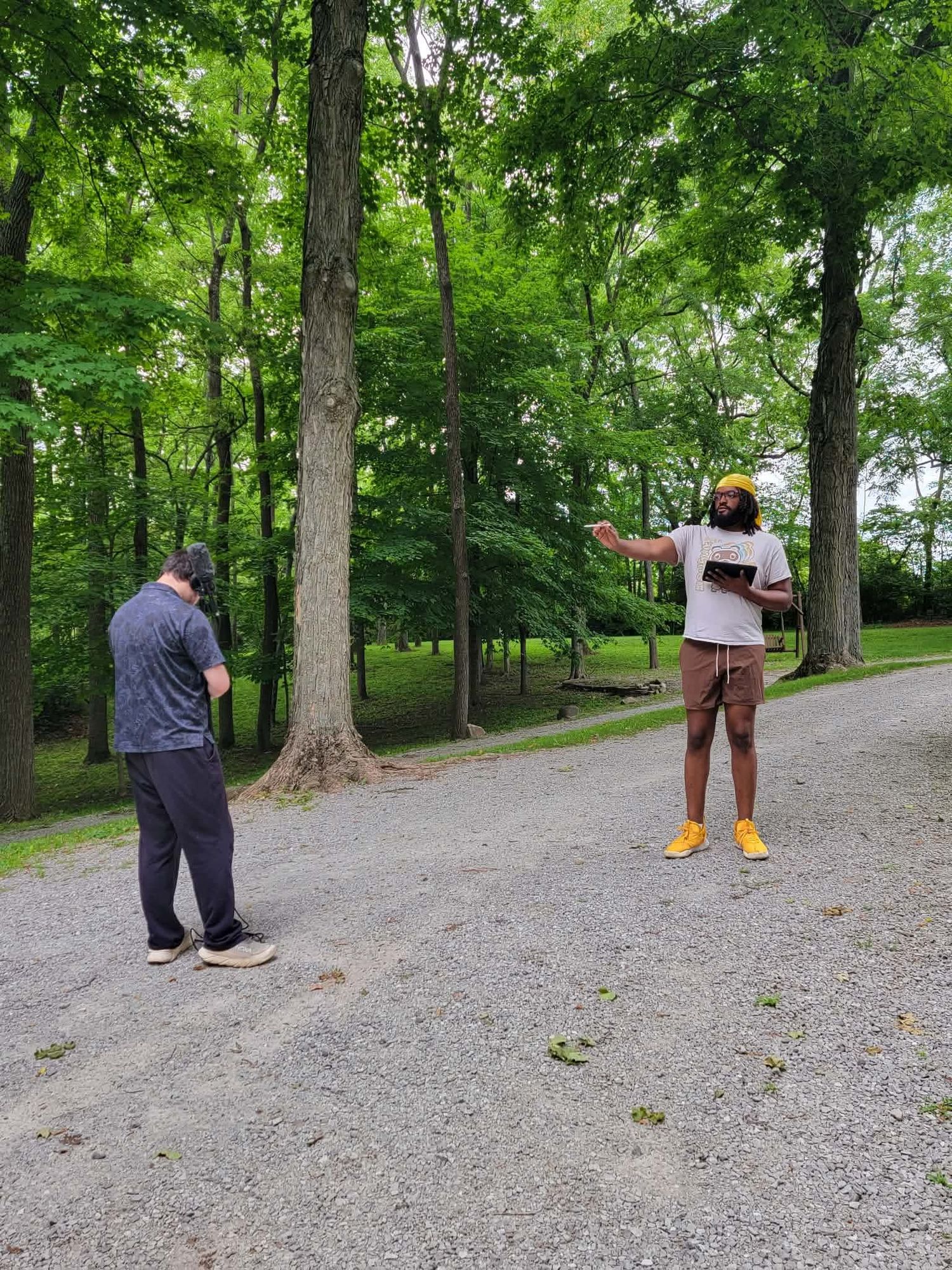 Two men stand on a gravel path in a park, one gesturing while holding a tablet outdoors.