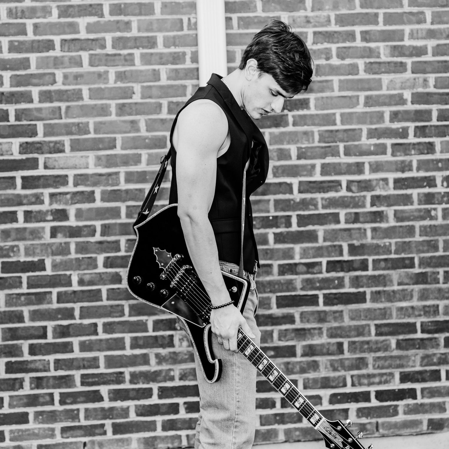 Young male guitarist in sleeveless shirt holding electric guitar against brick wall, black and white photo.