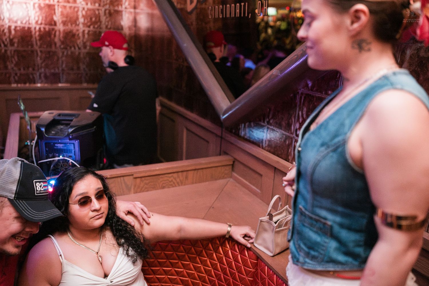 Two women conversing in a dimly lit bar booth area with a DJ setup visible in background.