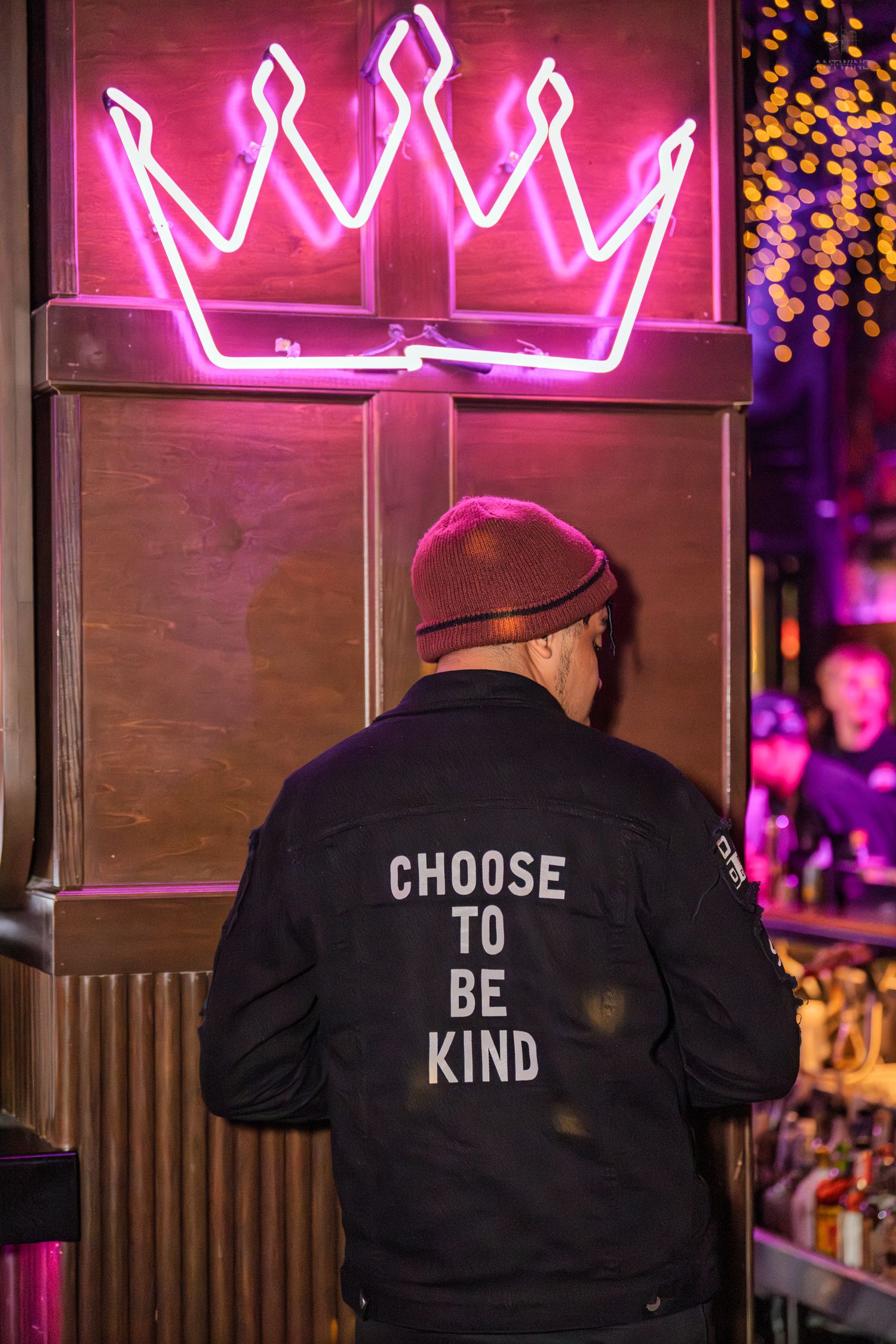 Person wearing a Choose To Be Kind jacket standing under a pink neon crown sign.