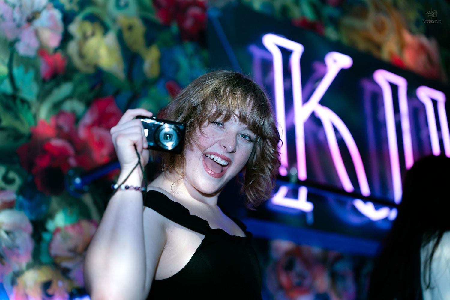 Smiling woman holding a small camera in front of a floral neon K sign.