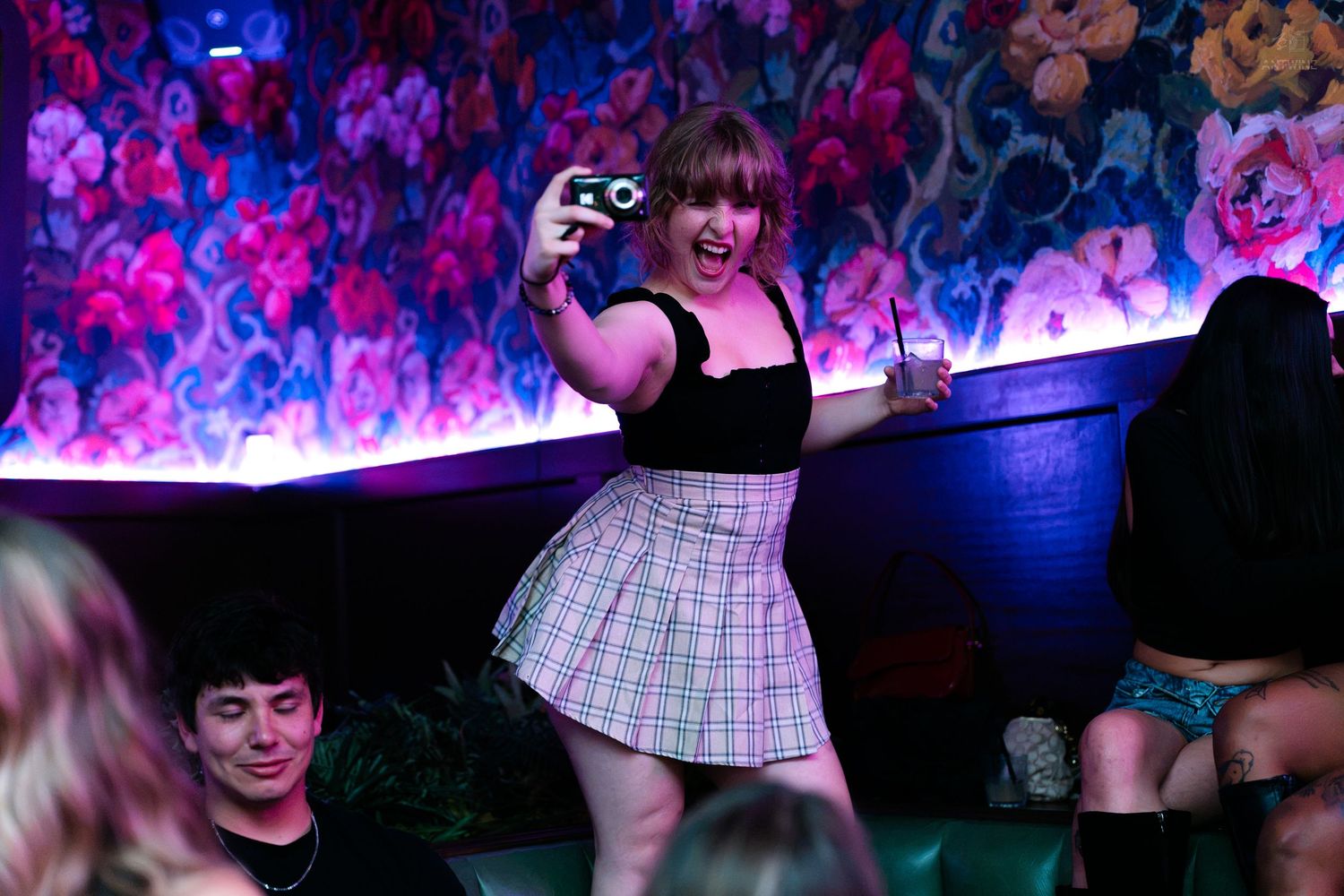 Woman in plaid skirt taking a photo with a camera at a nightclub.