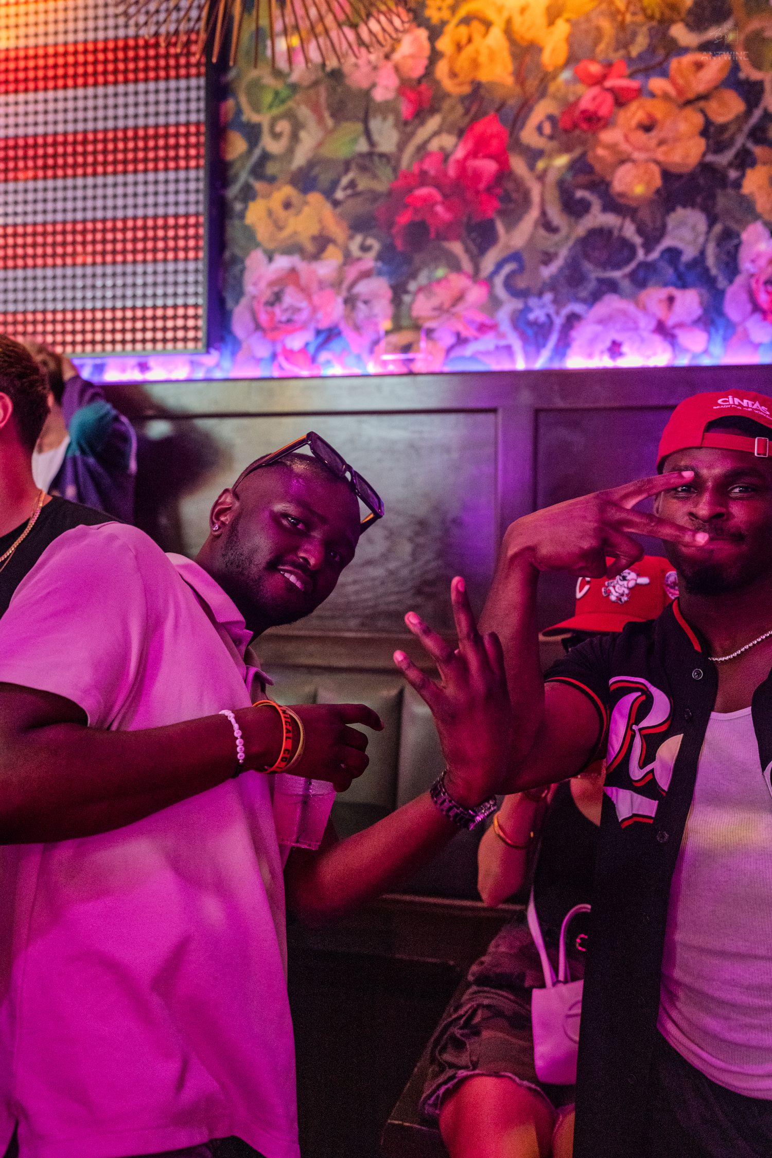 Three men posing with hand gestures in colorful neon-lit nightclub with floral wall mural.