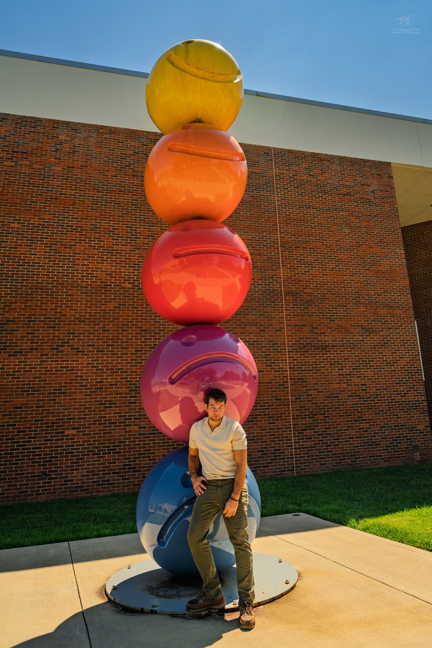 Man in white shirt posing beside tall colorful stacked ball sculpture against brick wall.