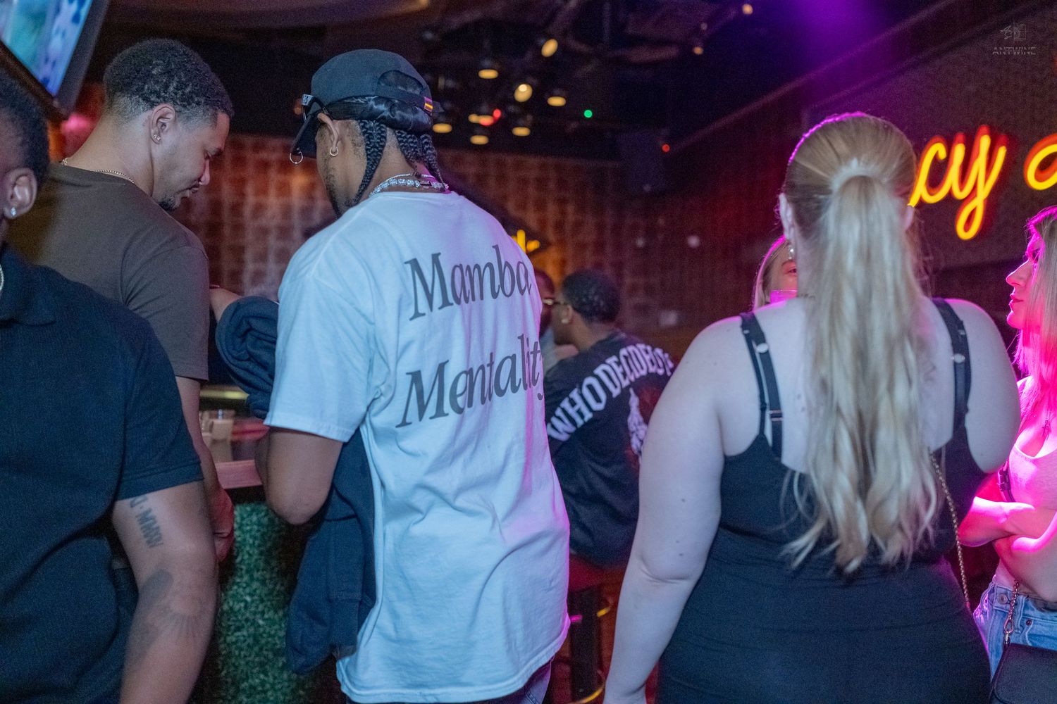Group of people socializing at a nightclub, one wearing a white Marbo Martali jersey shirt.