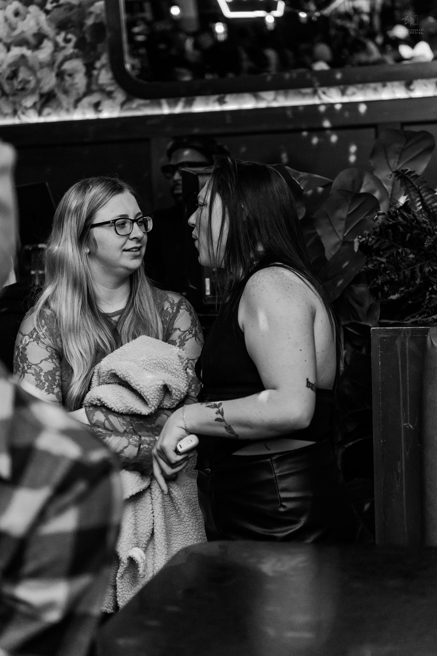 Two women conversing at a nightclub, one holding a blanket and wearing glasses.