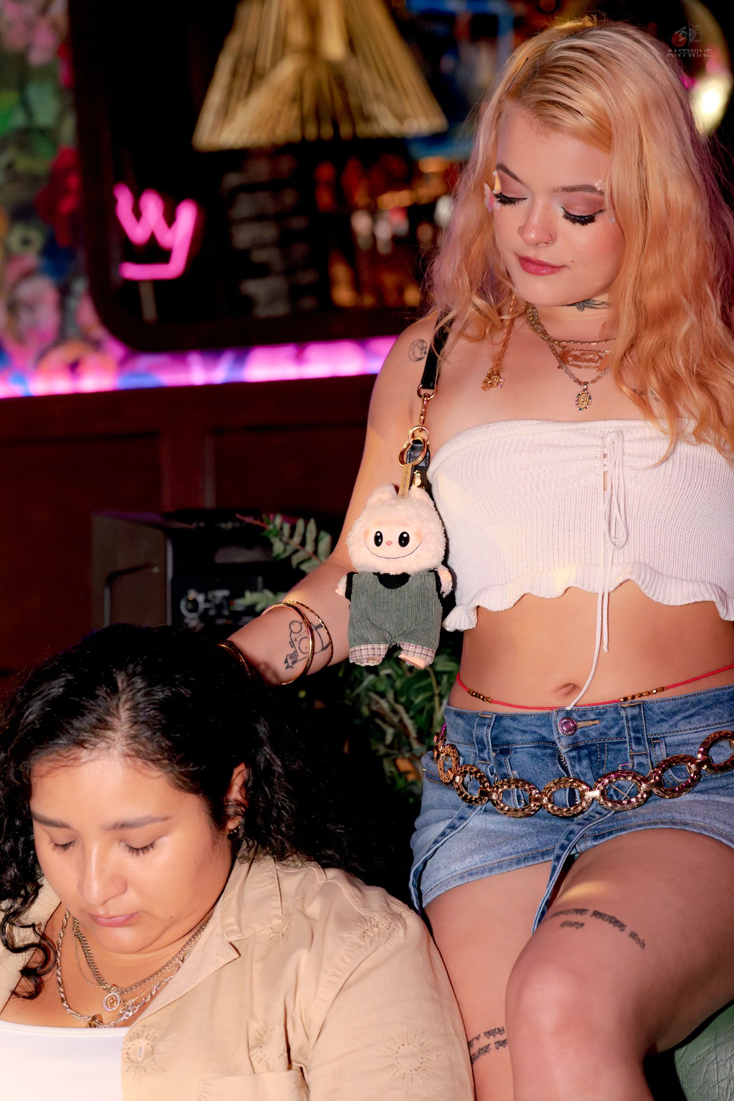 Two tattooed women in vibrant neon-lit bar, blonde wearing white crop top with toy charm.