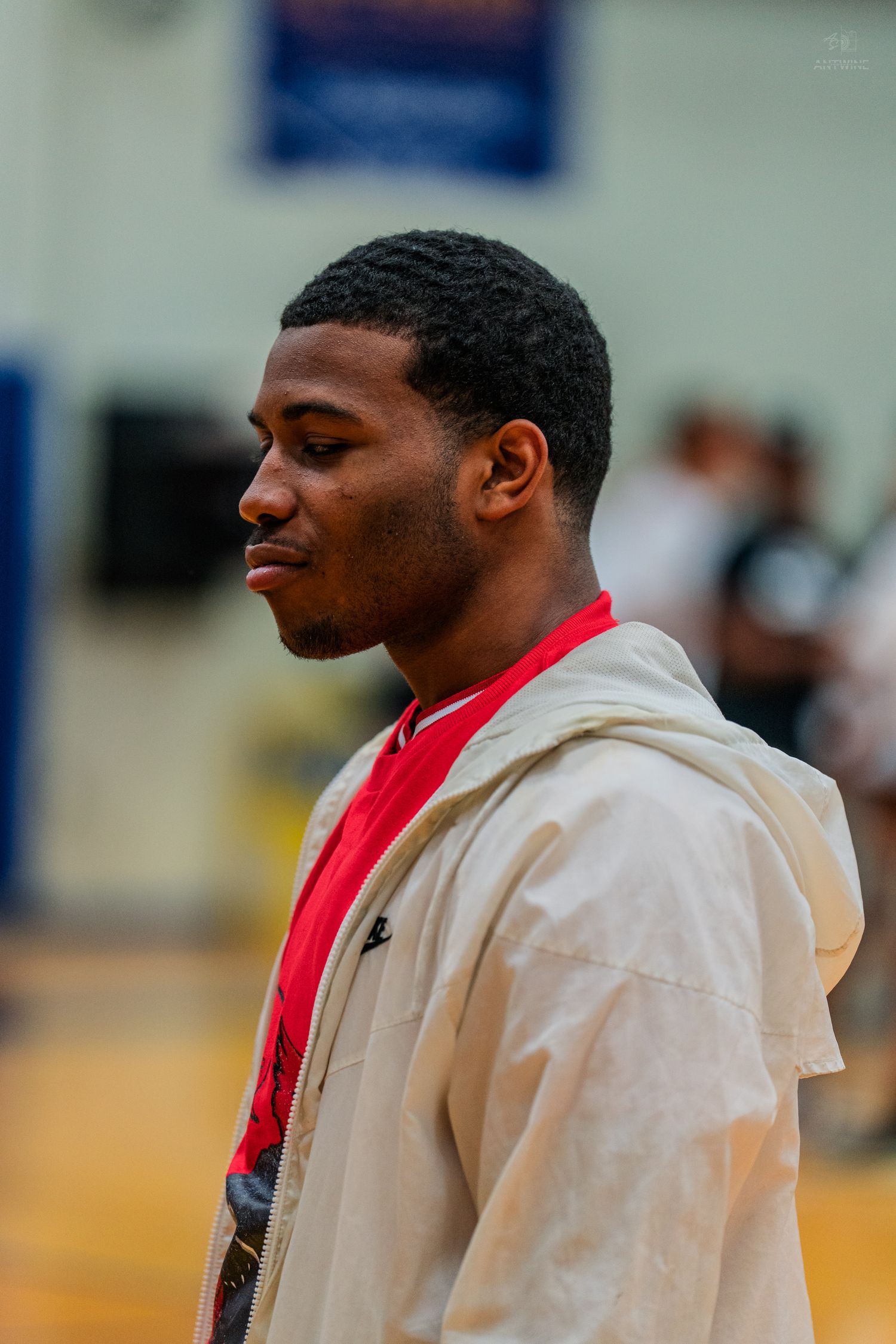Young Black man in beige hoodie and red shirt stands in a gym, looking sideways with a calm expression.