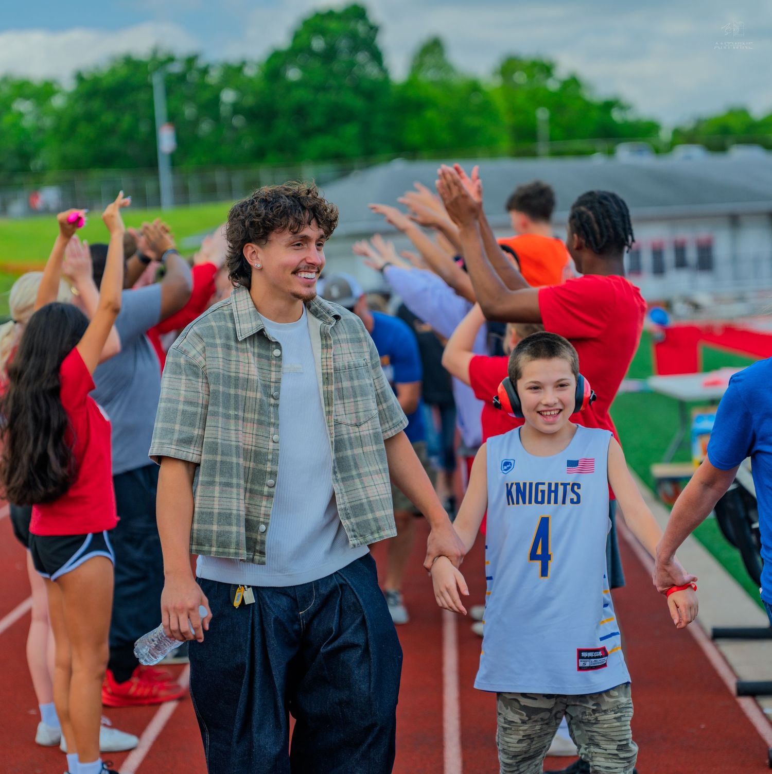 Two people holding hands on a track at a school sports event, one wearing a Knights jersey number 4, with cheering crowd behind.