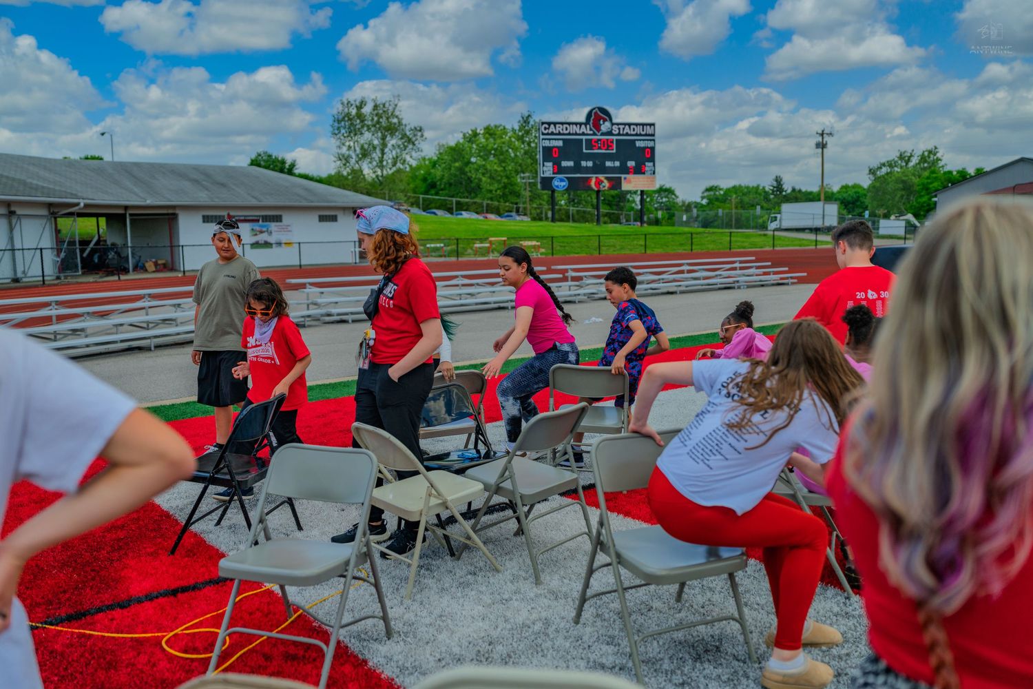 Group of people in red and pink shirts doing outdoor activities near metal chairs at a sports track on a sunny day.