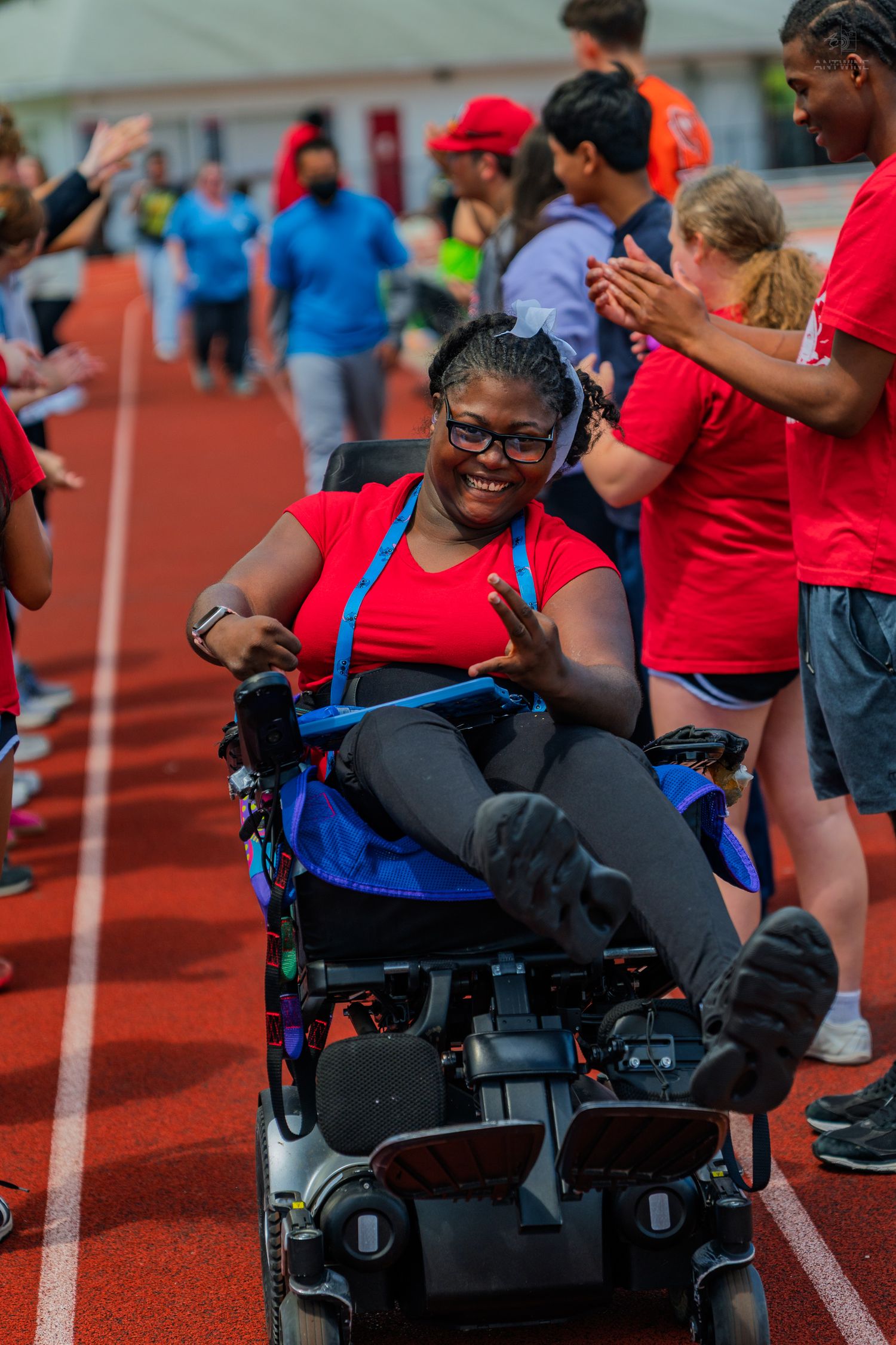Smiling woman in red shirt using a power wheelchair on a track, surrounded by cheering people at a Special Olympics event.