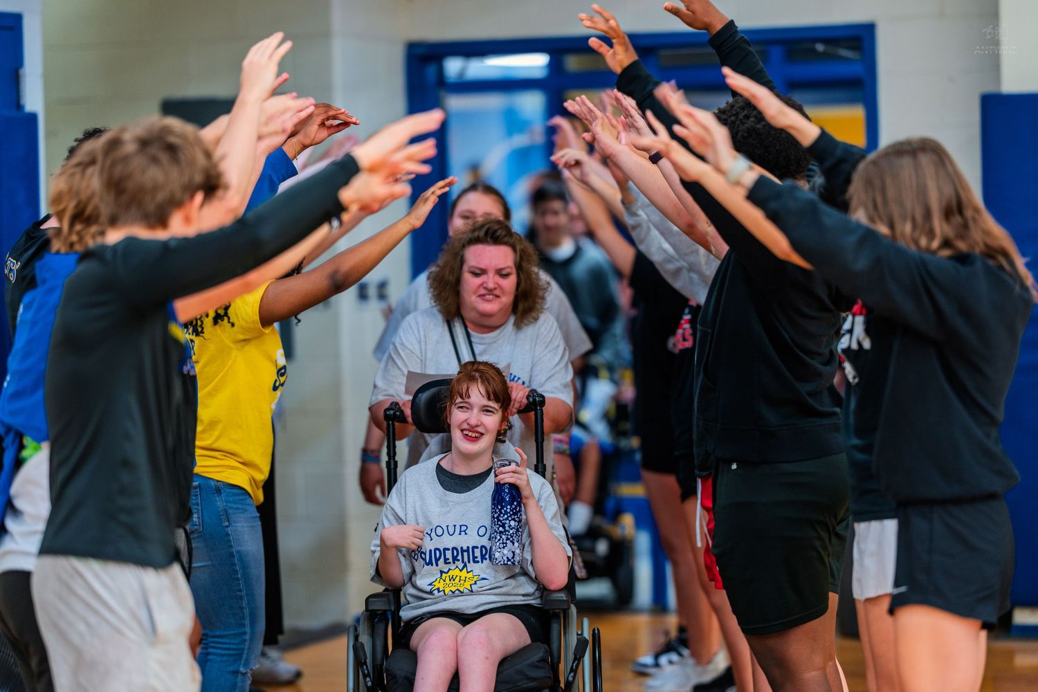 Child in wheelchair celebrated by cheering crowd forming arch in gymnasium, joyful special needs event.