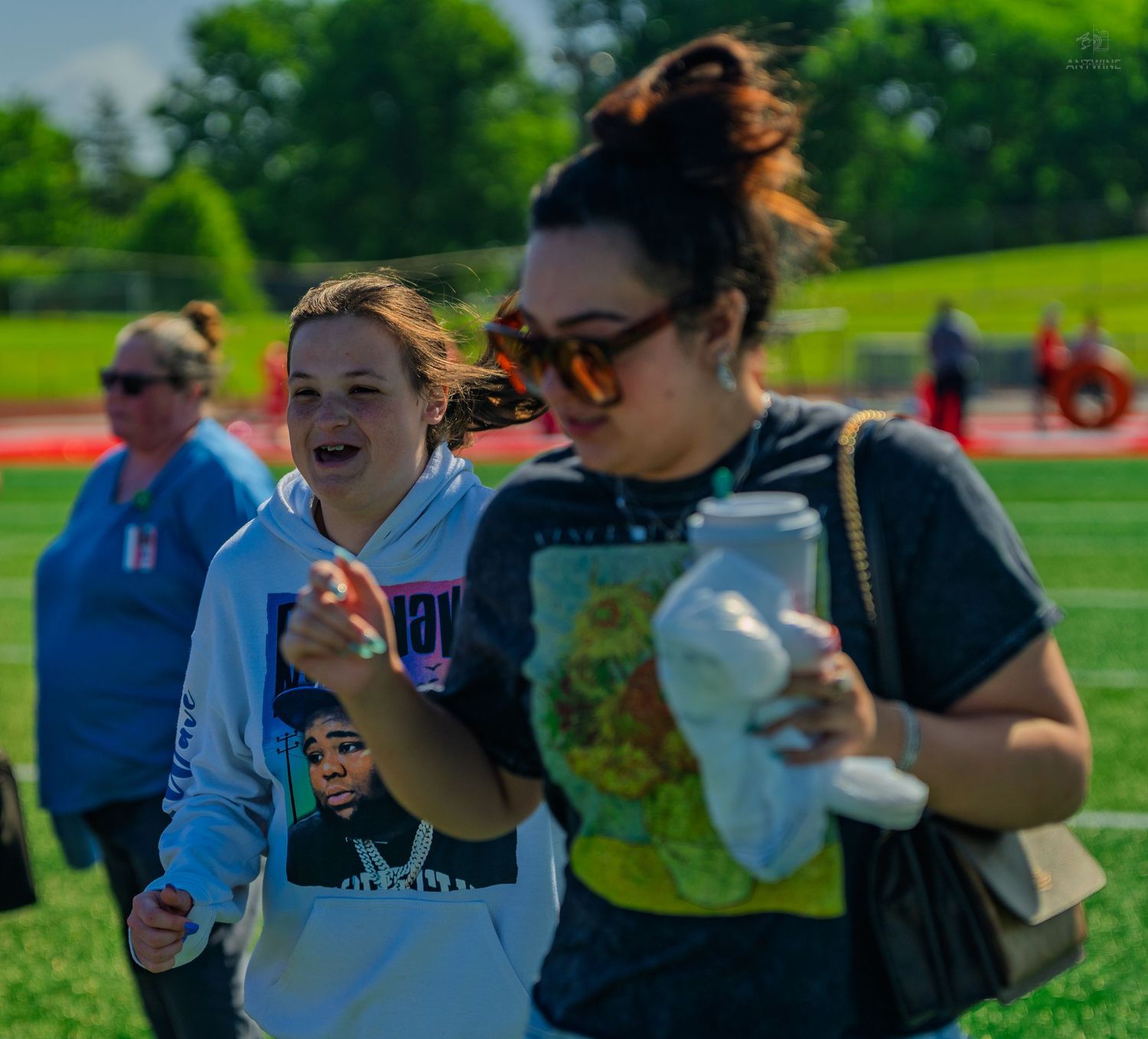 Two women walking on a sports field on a sunny day, one laughing in a hoodie, the other wearing sunglasses.