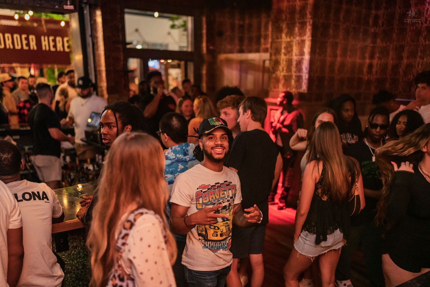 Crowded nightclub scene with diverse group of smiling people socializing near an Order Here sign.