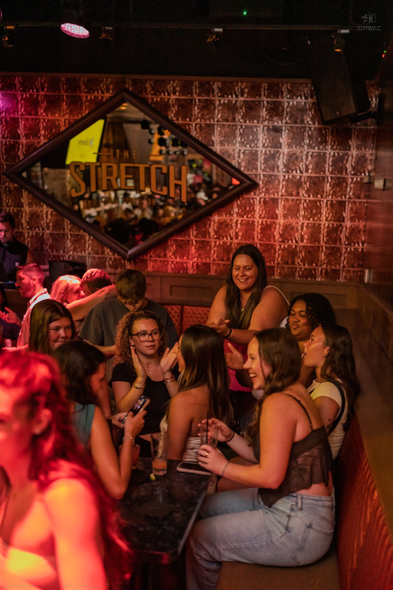 Group of women laughing and socializing at a bar under a Stretch nightclub sign with warm red lighting.