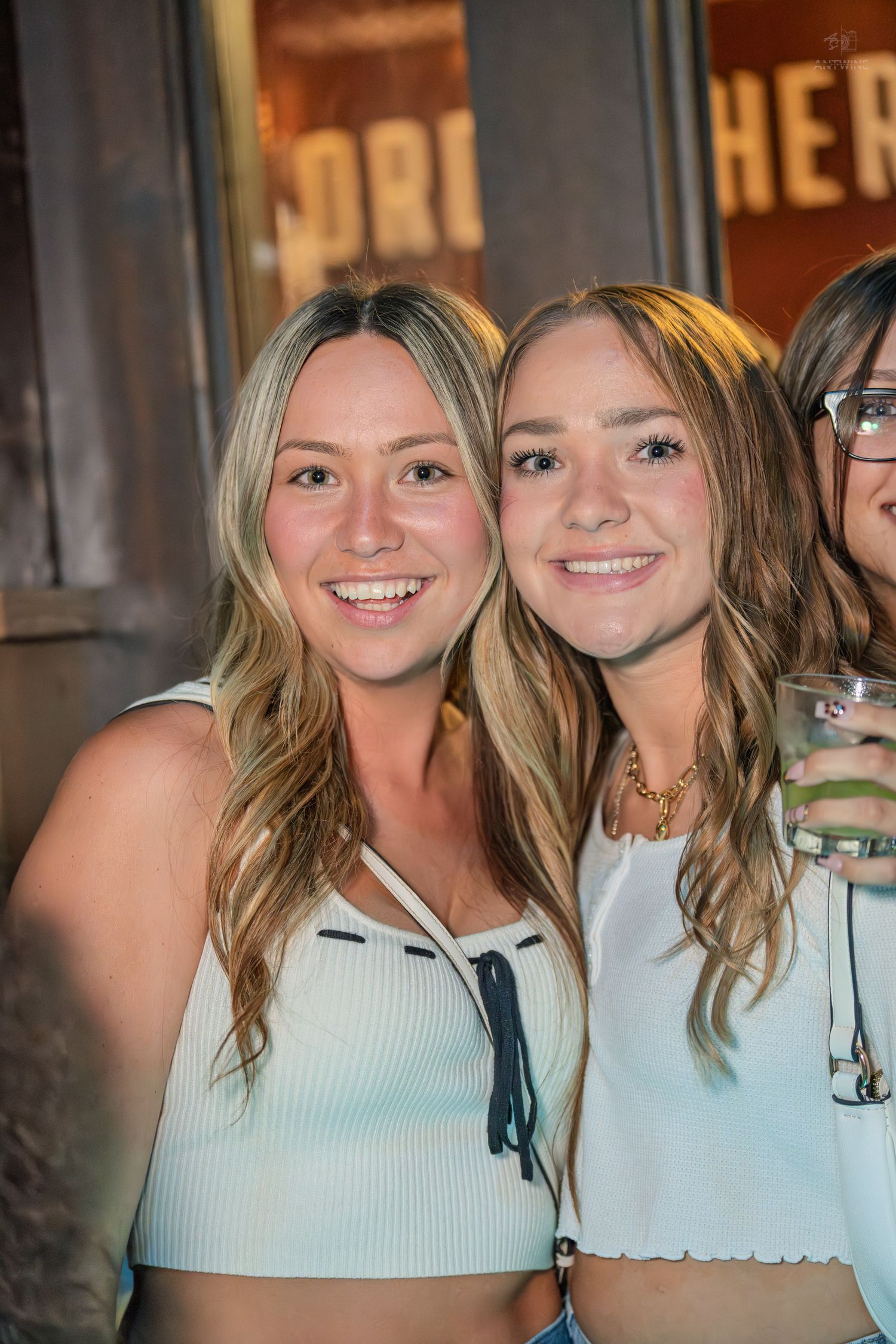 Two smiling blonde women in white tops pose together outside a bar or restaurant.