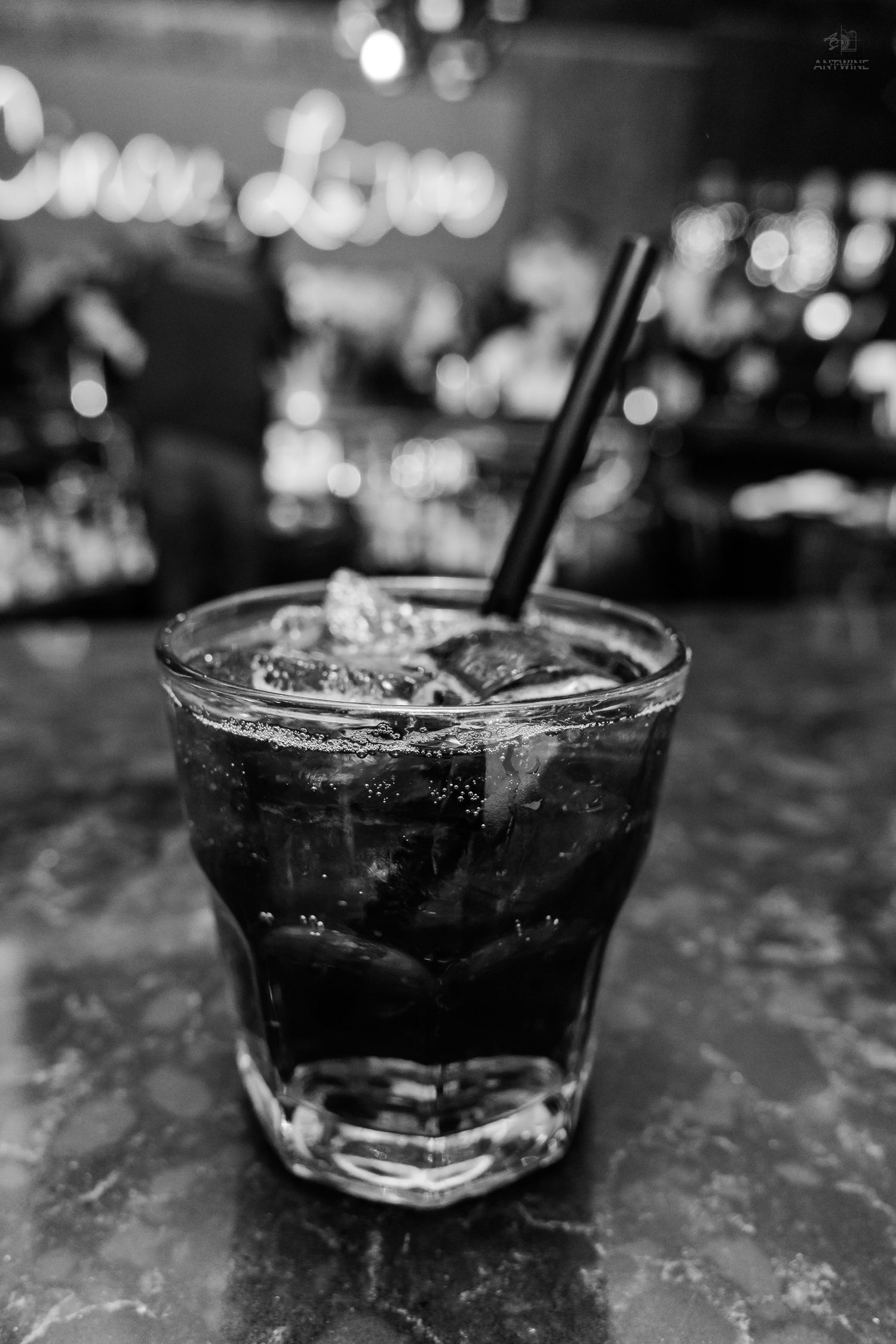 Black and white photo of a dark cocktail with ice and straws in a rocks glass on a marble bar.