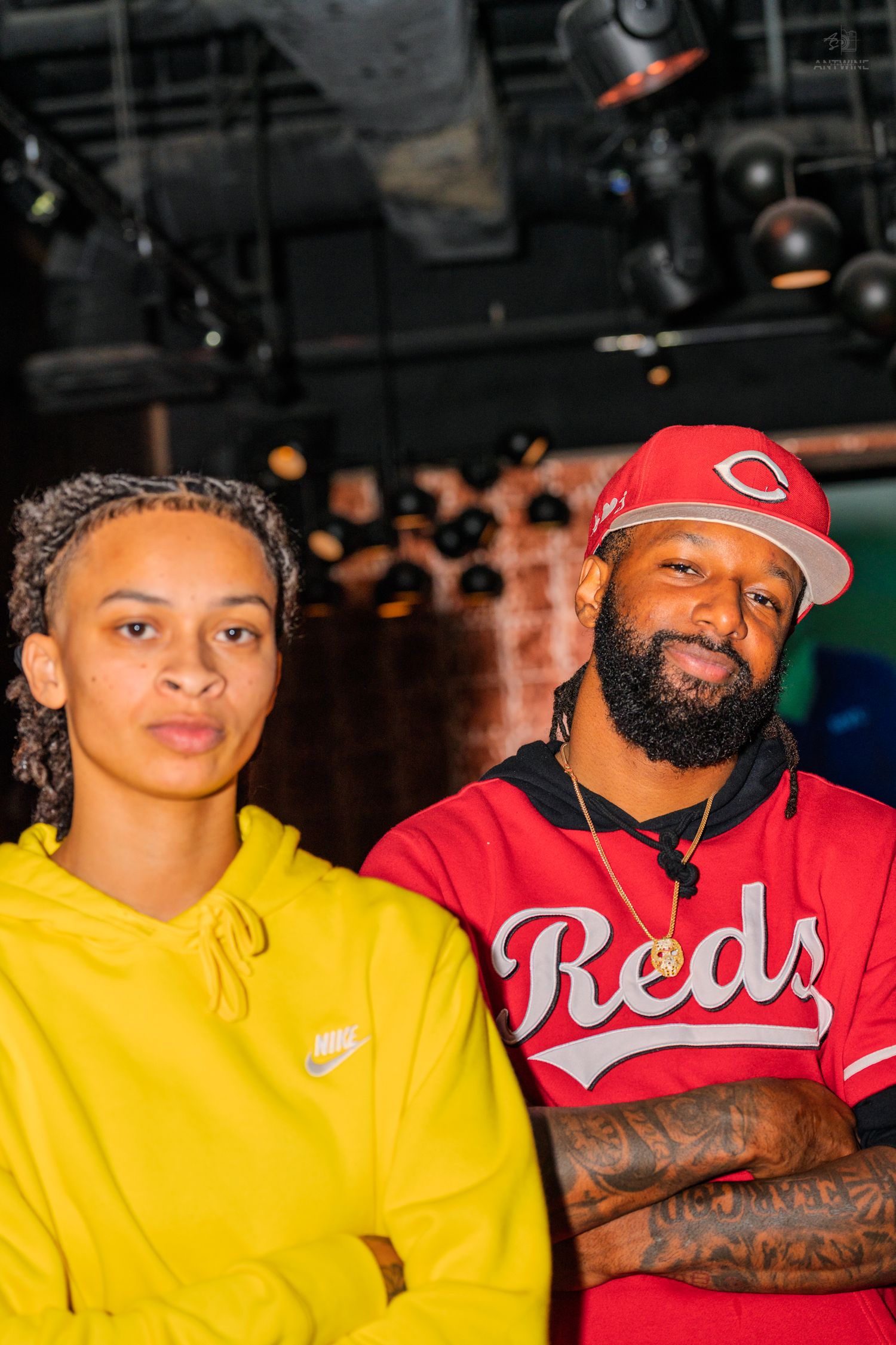Man in red Reds baseball jersey and woman in yellow Nike hoodie posing seriously at indoor venue.