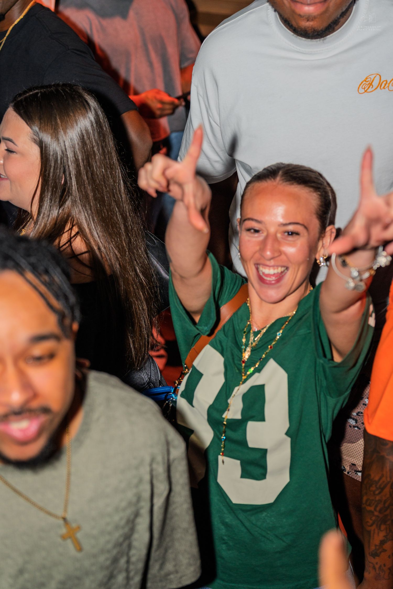 Woman in green football jersey smiling and gesturing at a crowded party.