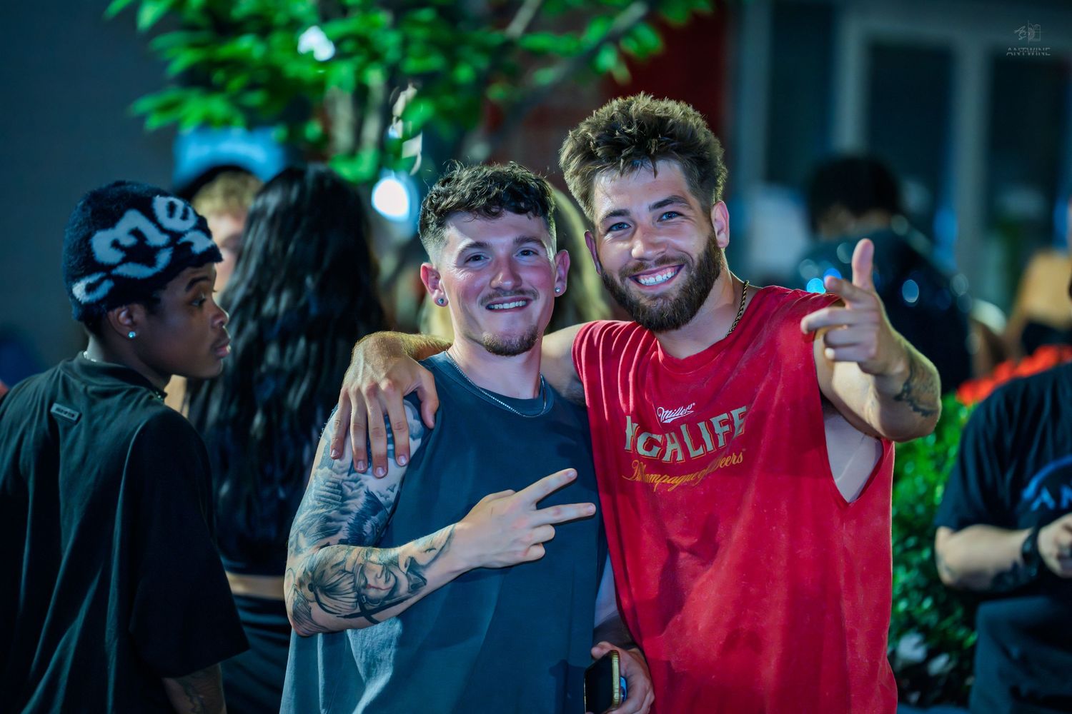Two smiling young men pose together outdoors at night, one flashing a peace sign.
