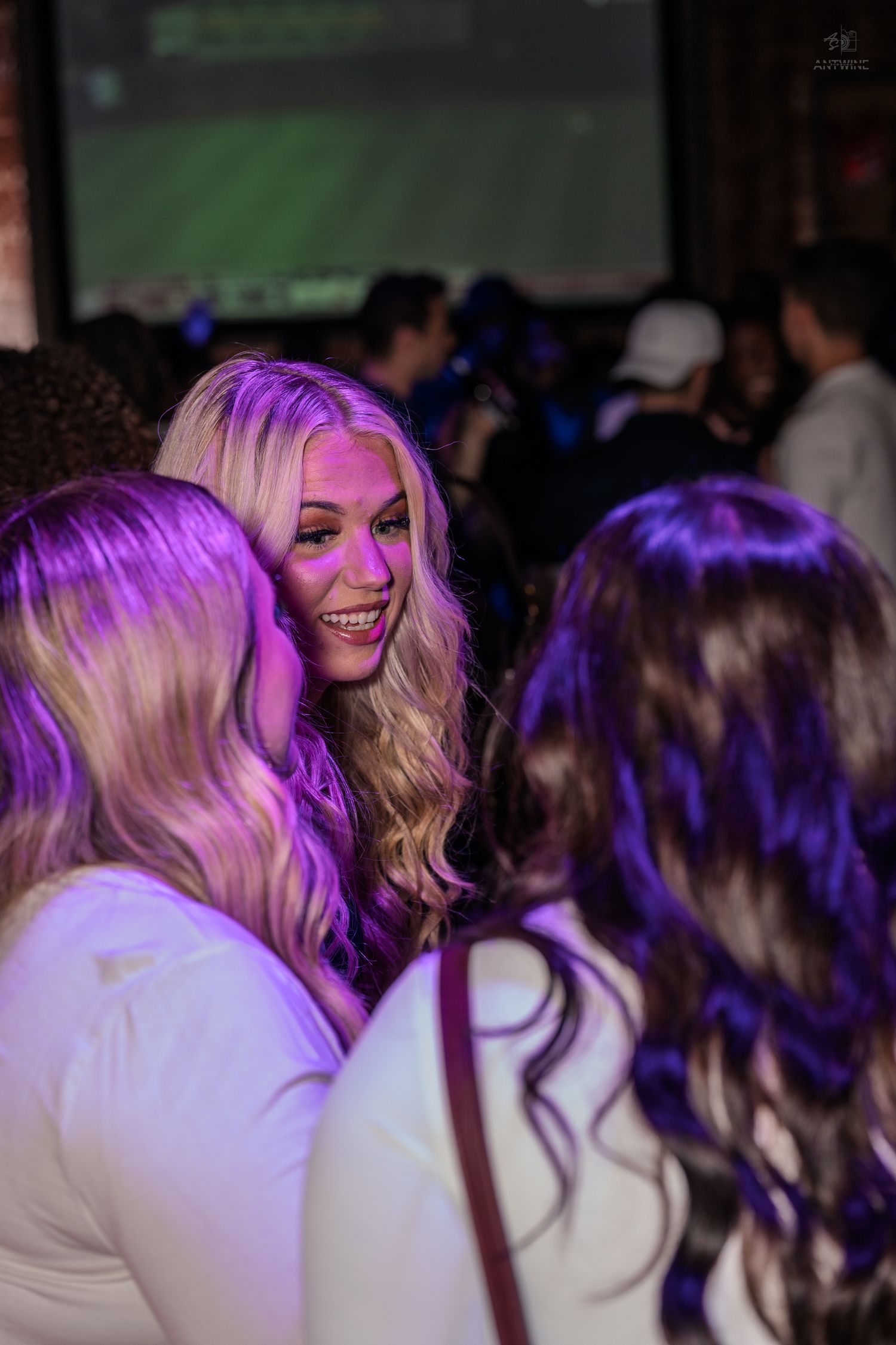 Smiling blonde woman in white top socializes with friends under purple lighting at a crowded nightclub.