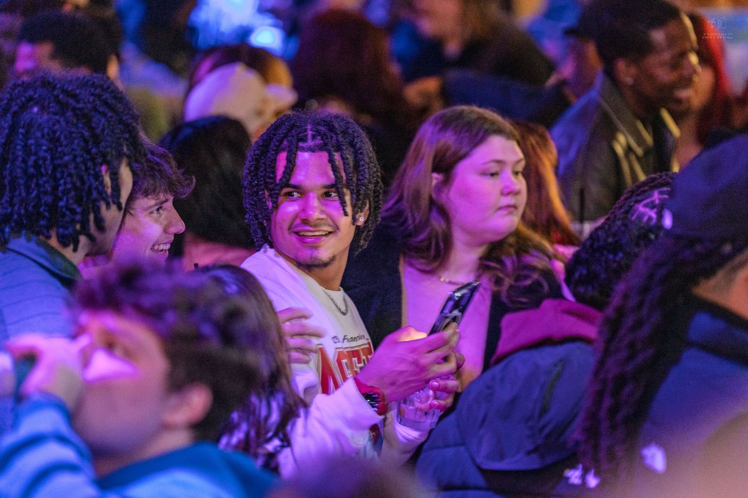 Group of young people smile and chat at a colorful indoor concert under purple lights.