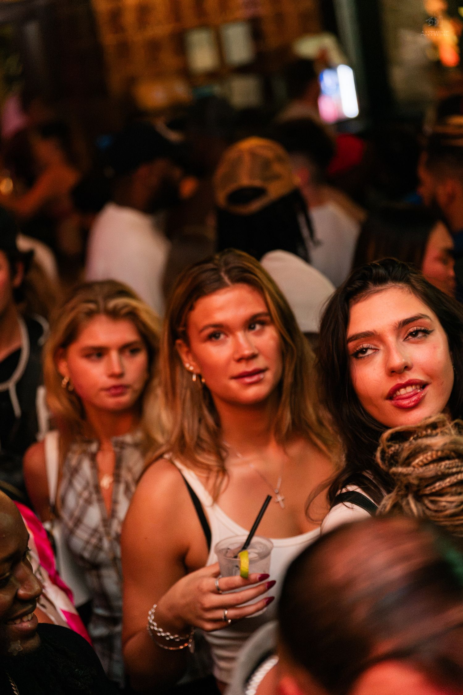 Three young women socializing at a crowded nightclub, one holding a cocktail.