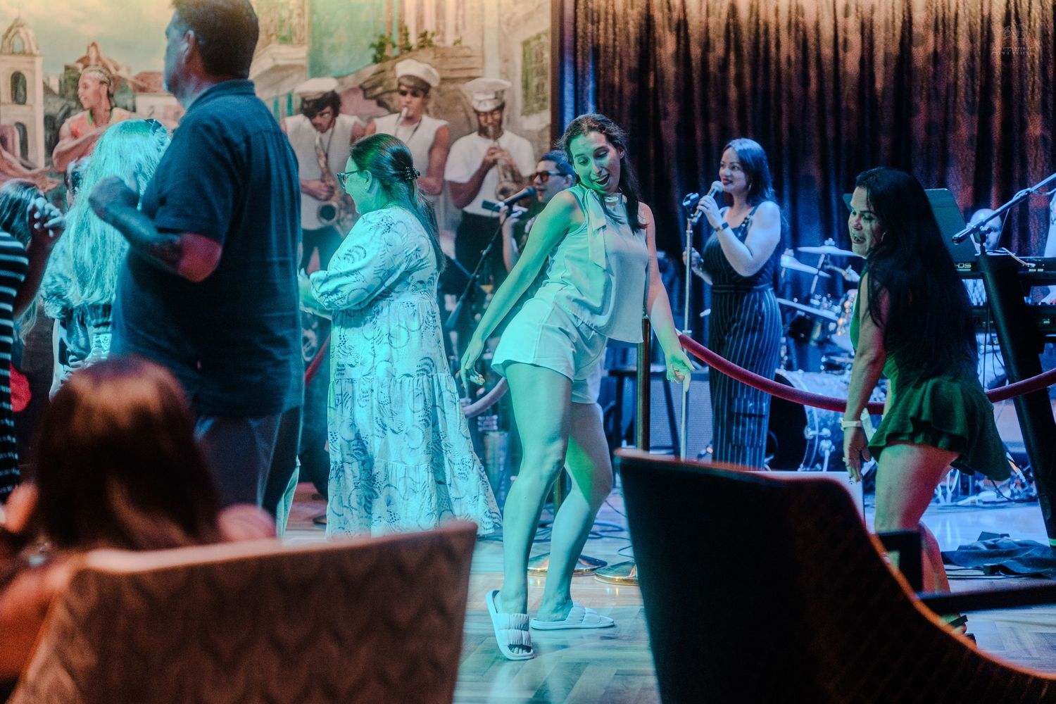 Energetic live band performance with dancers in white dresses on a cruise ship stage.