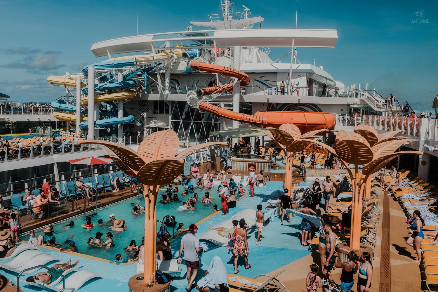 Crowded cruise ship pool deck with water slides, palm tree decorations, and passengers swimming.