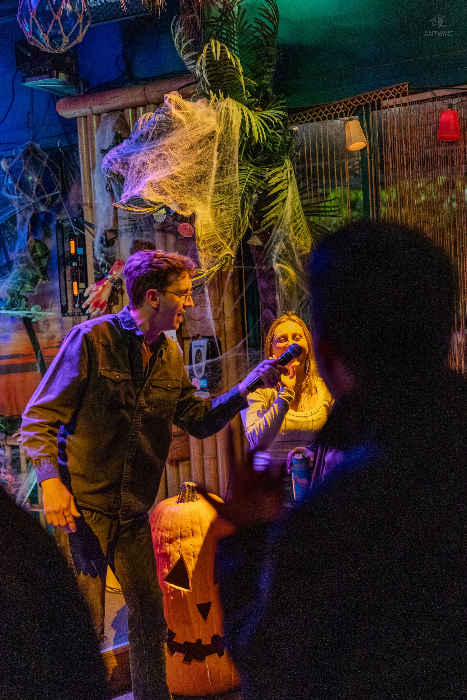 Man holds microphone to woman at Halloween-decorated karaoke bar with cobwebs and jack-o-lantern.