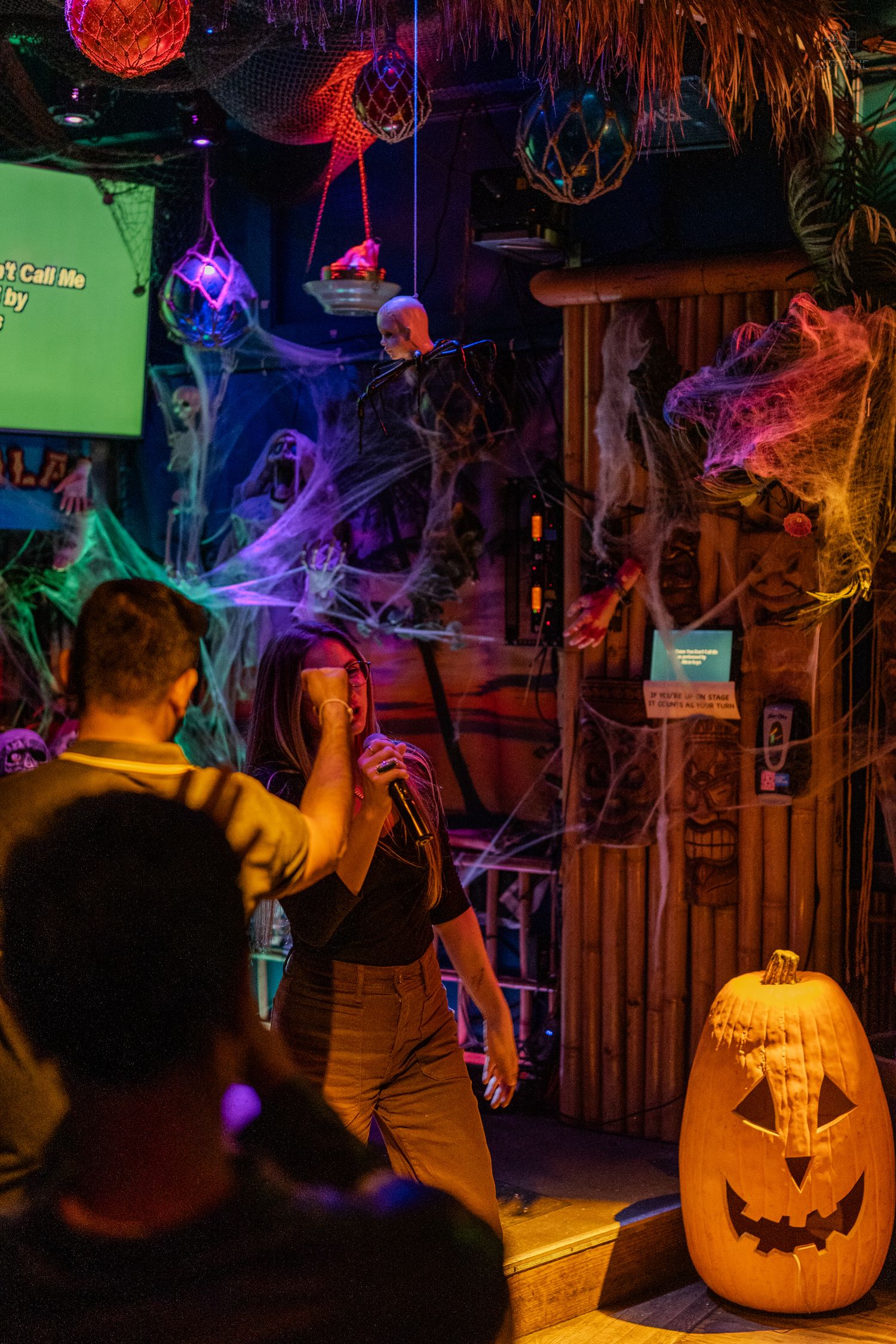 Halloween karaoke bar scene with glowing jack-o-lantern, colorful cobwebs, and performer on decorated stage.