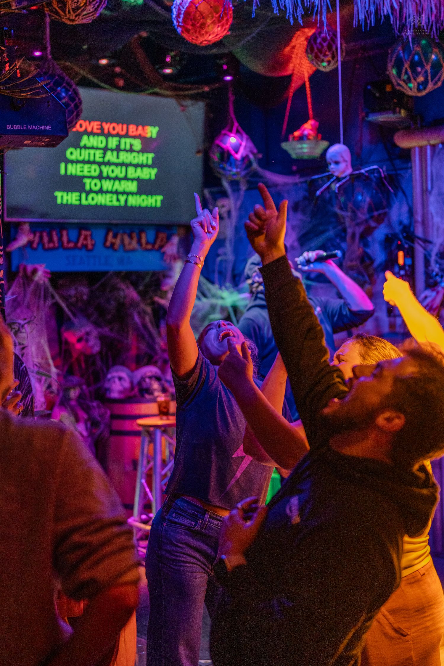 Excited crowd dancing and pointing at karaoke lyrics screen in a vibrant neon-lit bar.