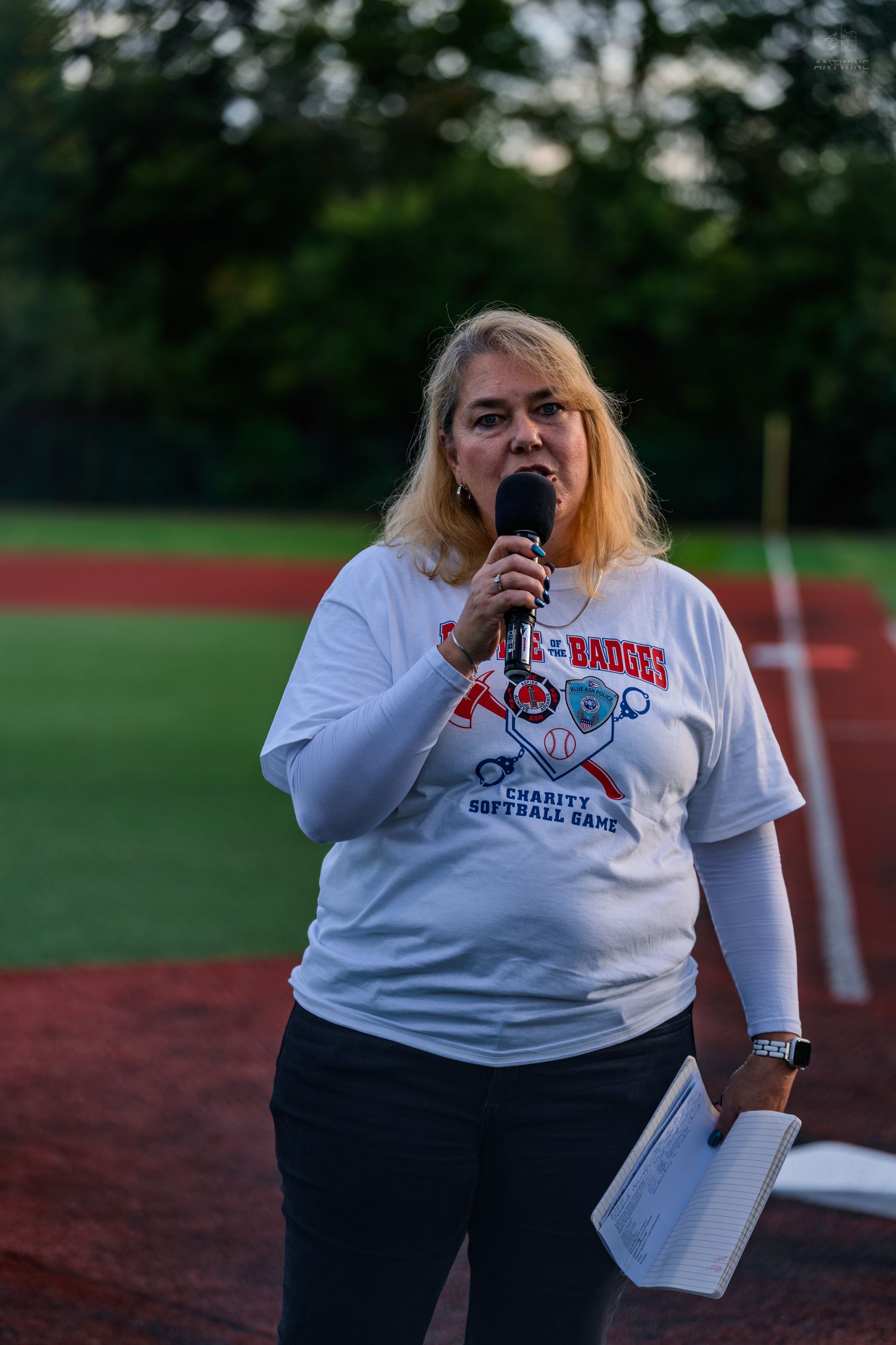 Woman in charity softball game shirt speaks into microphone at baseball field ceremony at dusk.