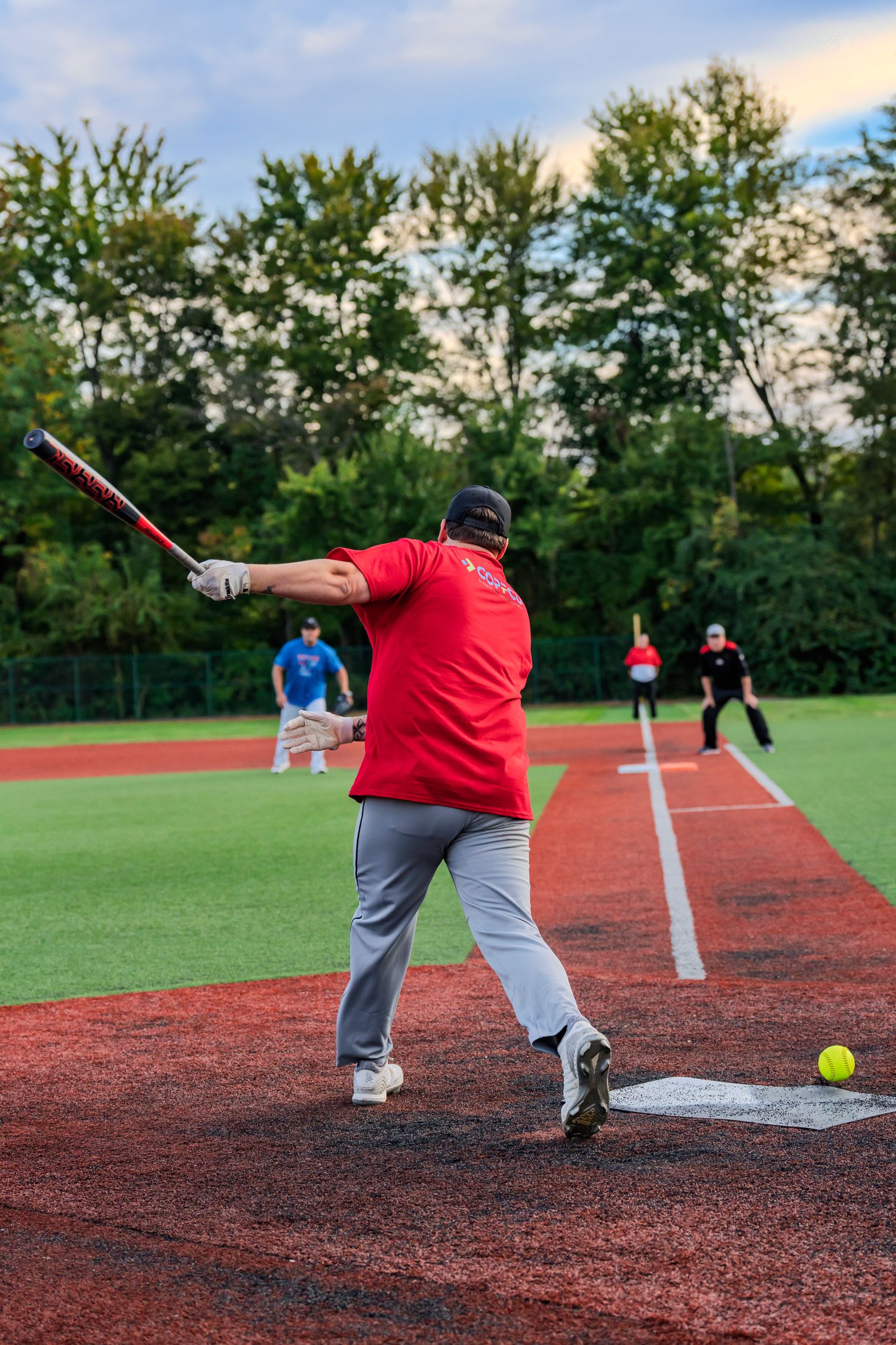 A batter in a red shirt swings at a yellow softball near home plate on a red and green baseball field.
