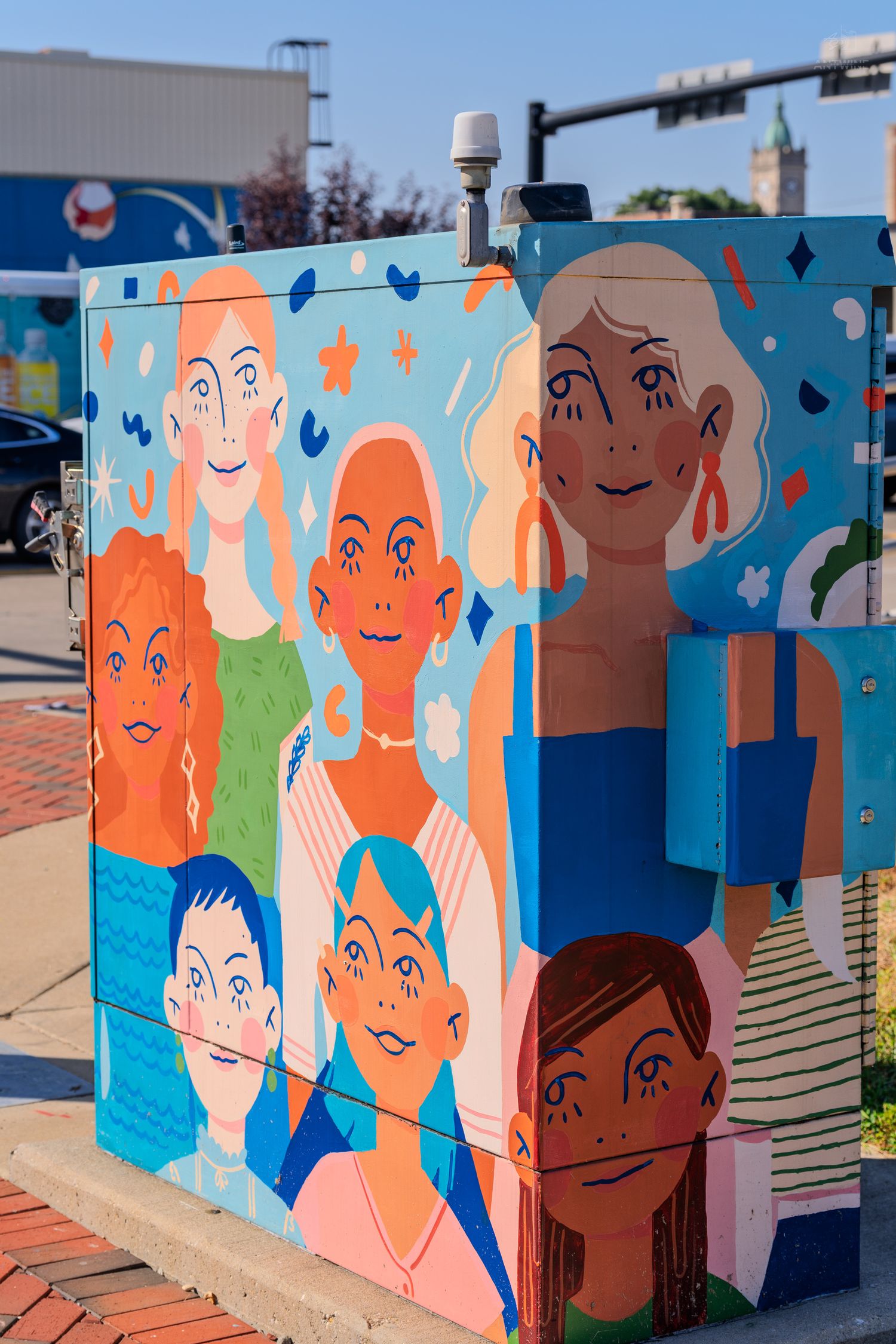 Colorful street art mural on utility box featuring diverse cartoon faces in vibrant blues, oranges, and greens.