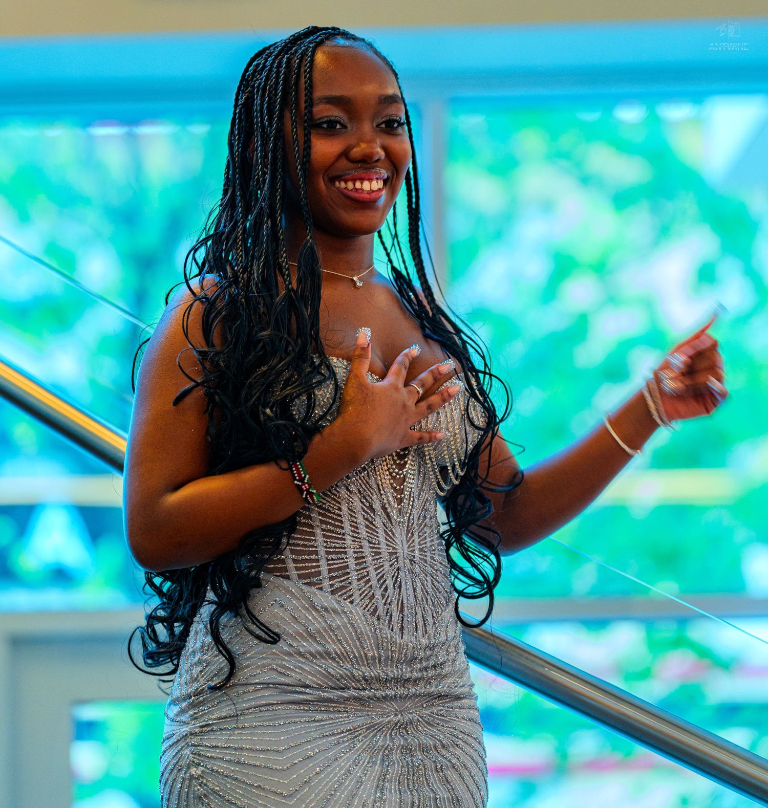 Young woman in elegant silver beaded gown with long braids smiling on staircase.