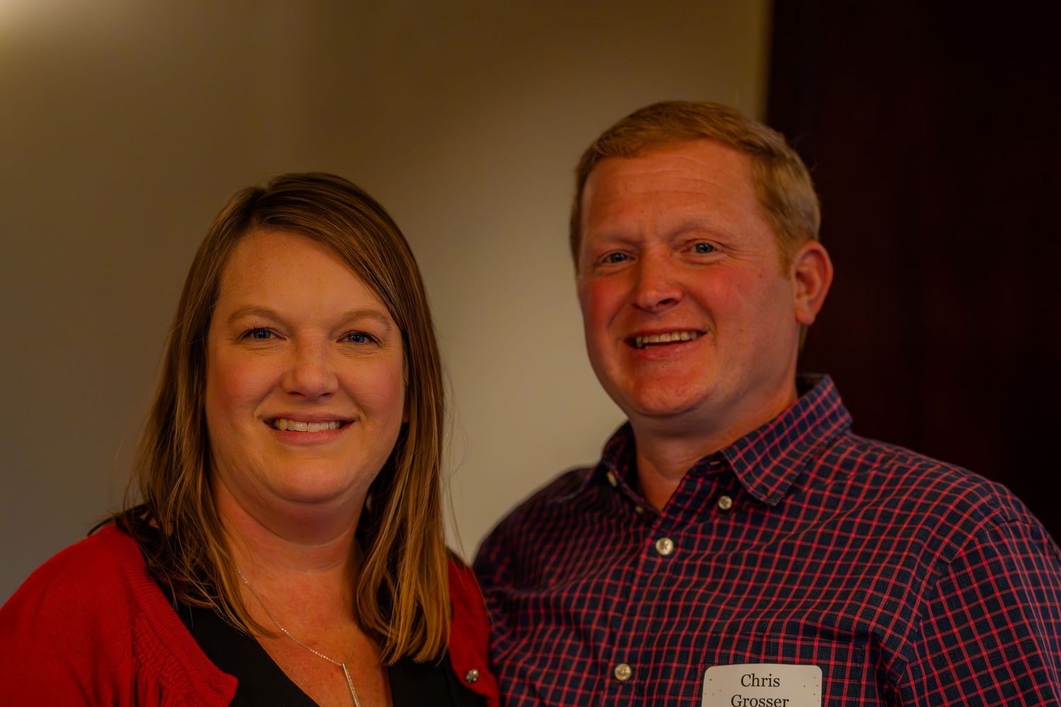 A smiling man and woman pose together at an indoor event, the man wearing a name tag and plaid shirt.