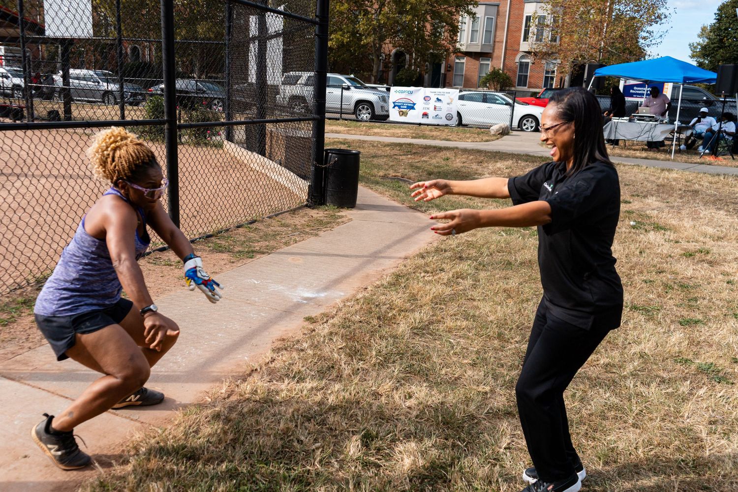 Two women playing catch outdoors near a chain-link fence at a sunny community event.