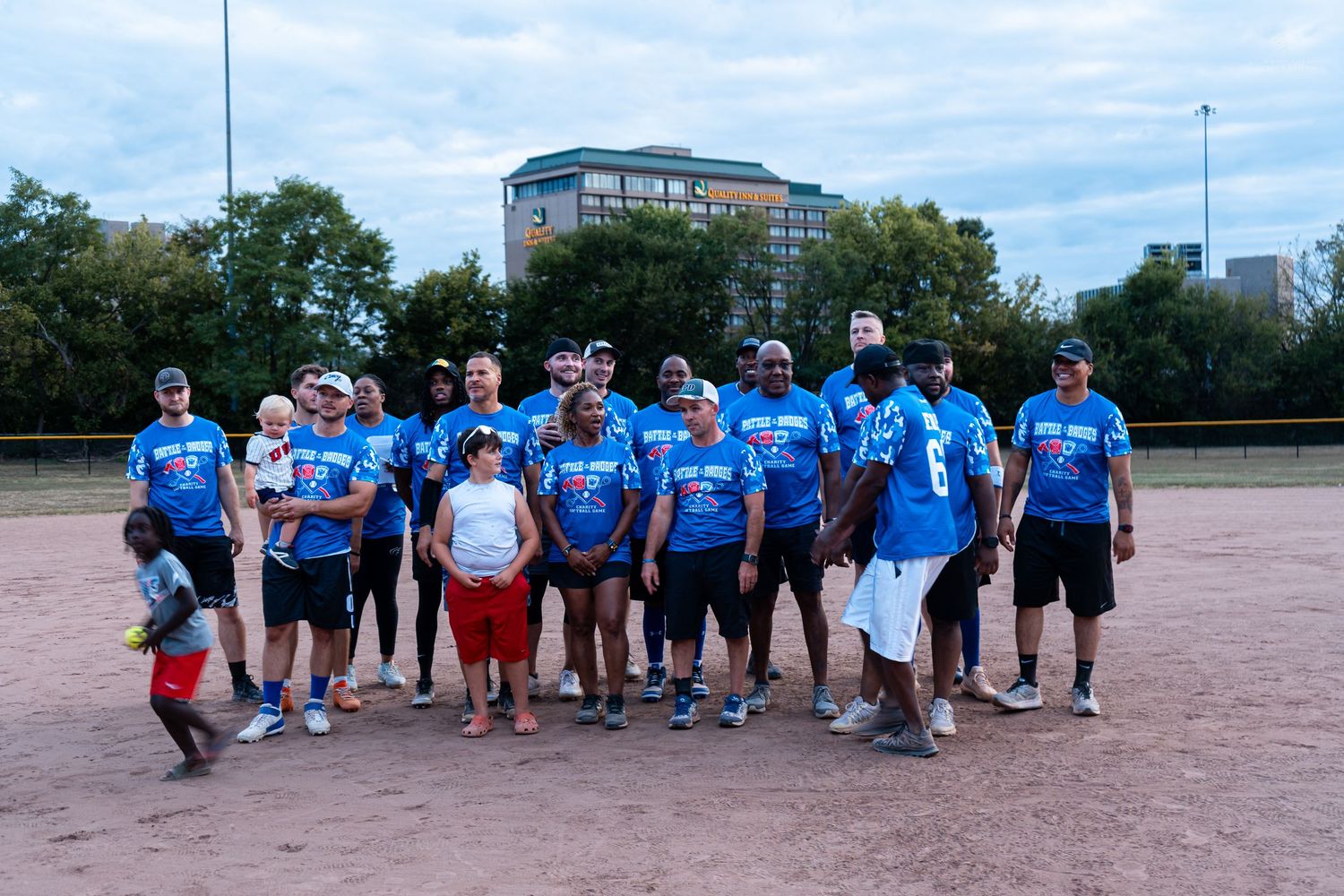 A group of softball players in blue COPFCU jerseys pose together on a dirt baseball field with trees in the background.