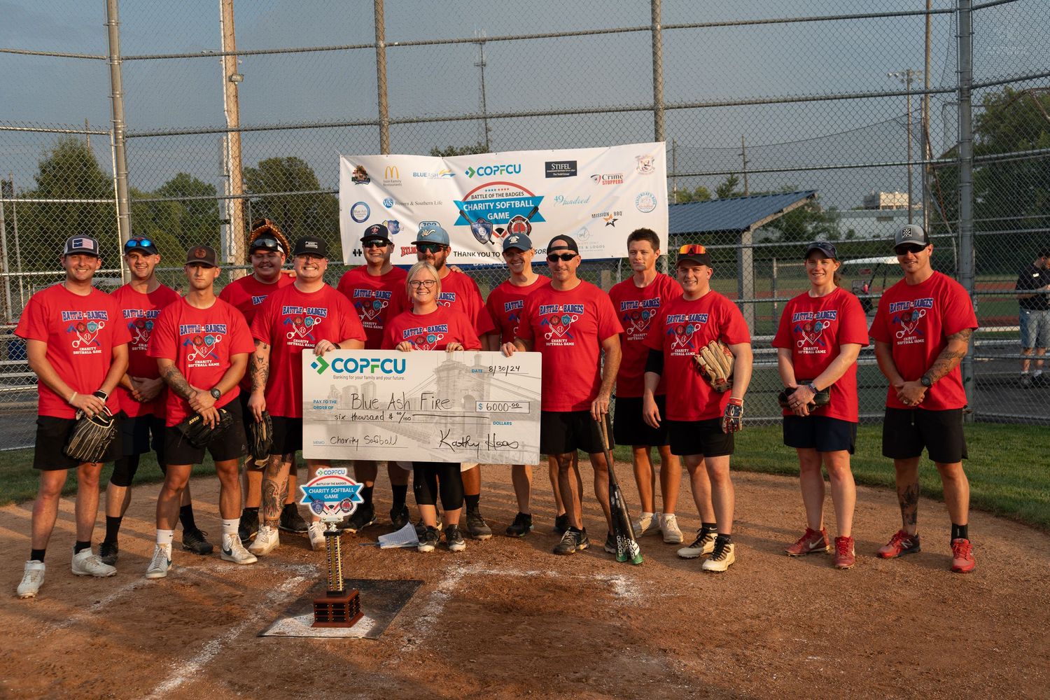 Softball team in red jerseys poses with trophy and oversized check on outdoor diamond after championship win.