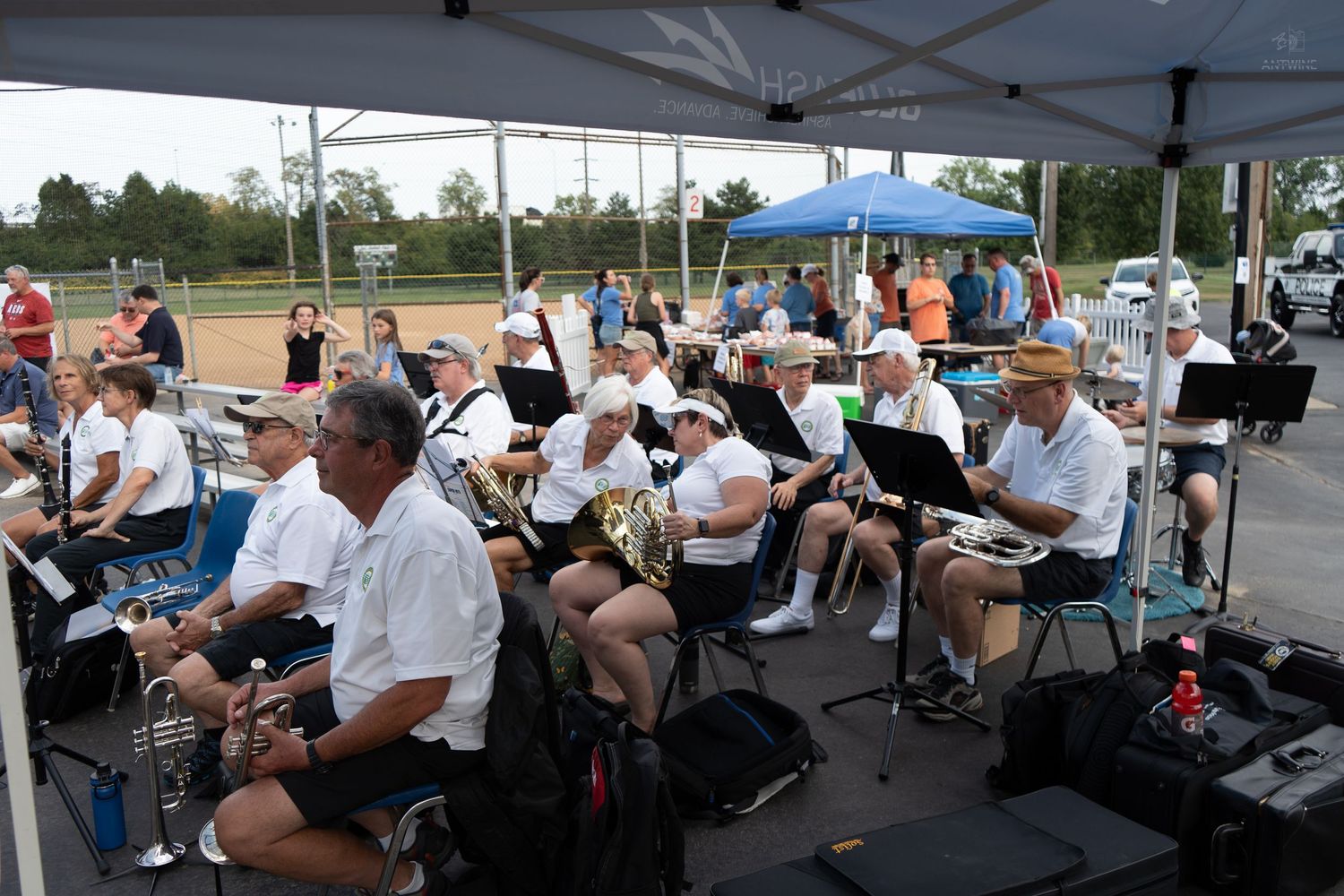 Outdoor band concert under a white tent with musicians in white shirts playing brass instruments at a sunny community event.